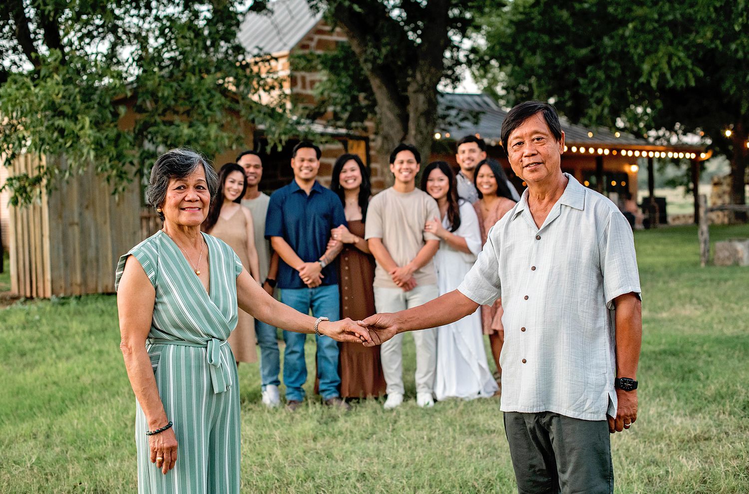 An elderly Asian couple holds hands in a backyard while their large family gathers behind them under a tree.