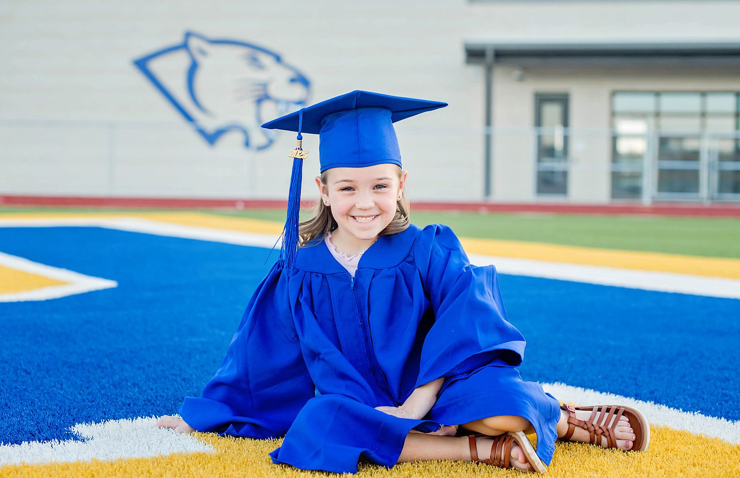 Graduate in bright blue cap and gown laying on blue and yellow sports turf with joyful expression.