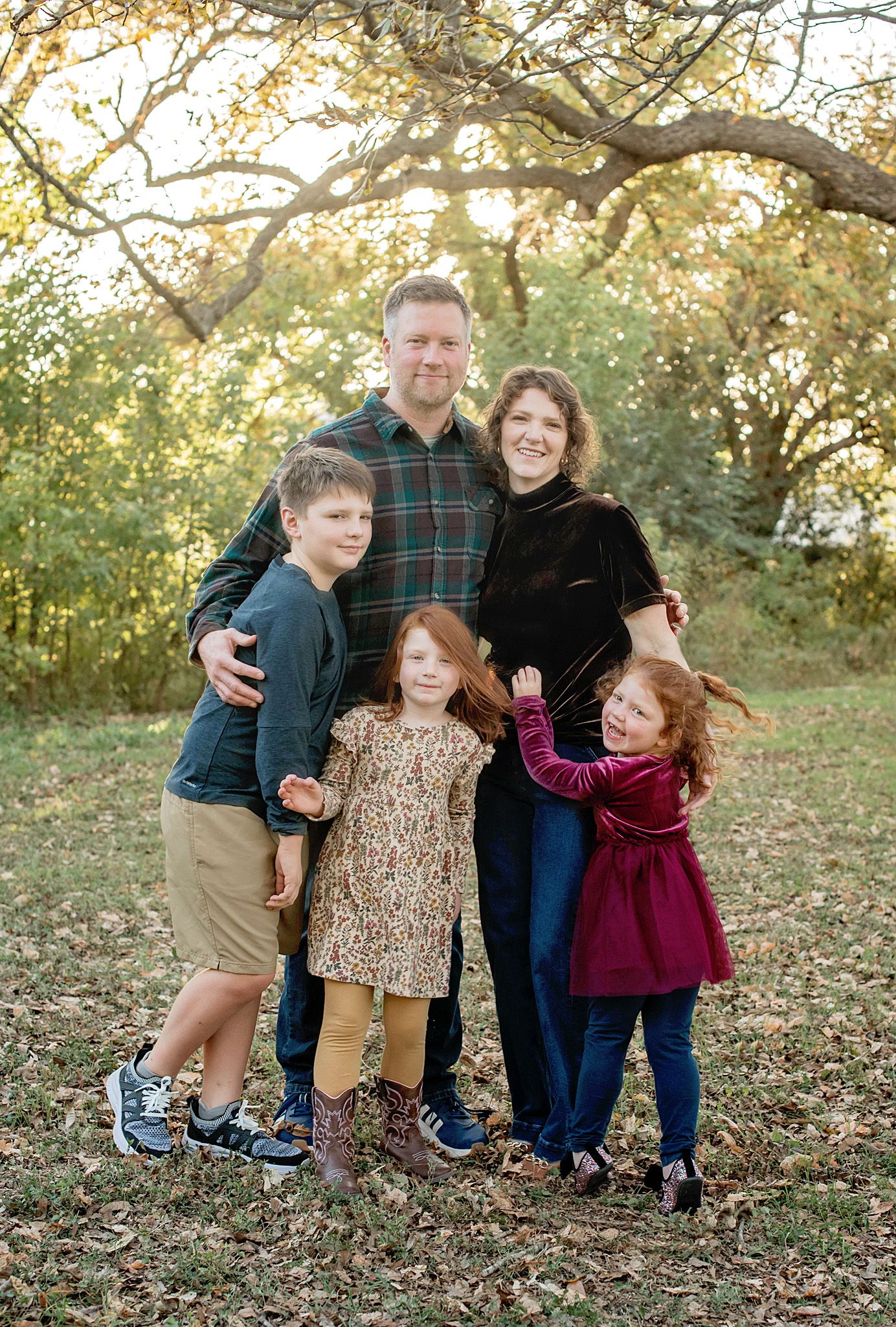 A family poses together outdoors on fallen autumn leaves with trees in the background during golden hour.