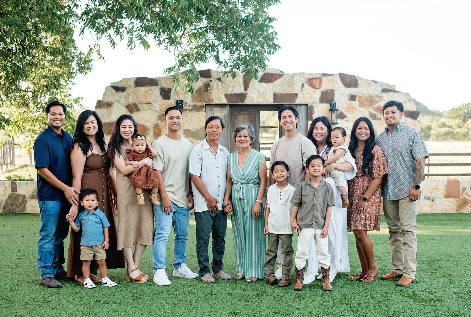 Large multigenerational Asian family posing together outdoors on green grass near a stone structure.