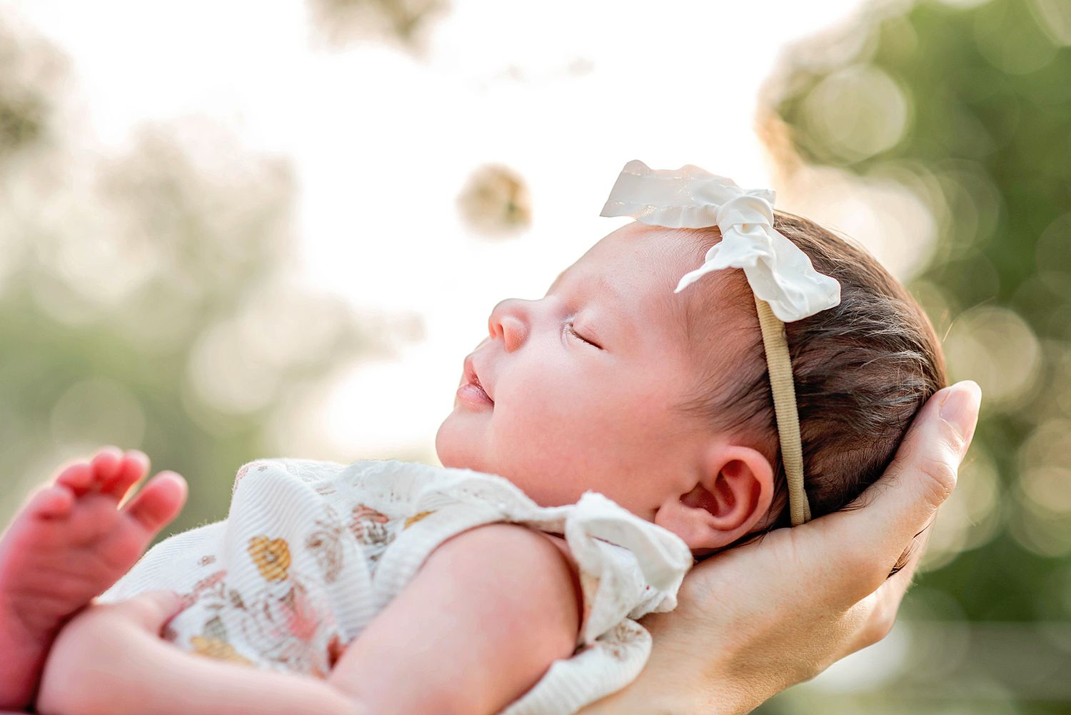 Close-up of a sleeping newborn baby wearing a white bow headband in gentle sunlight.