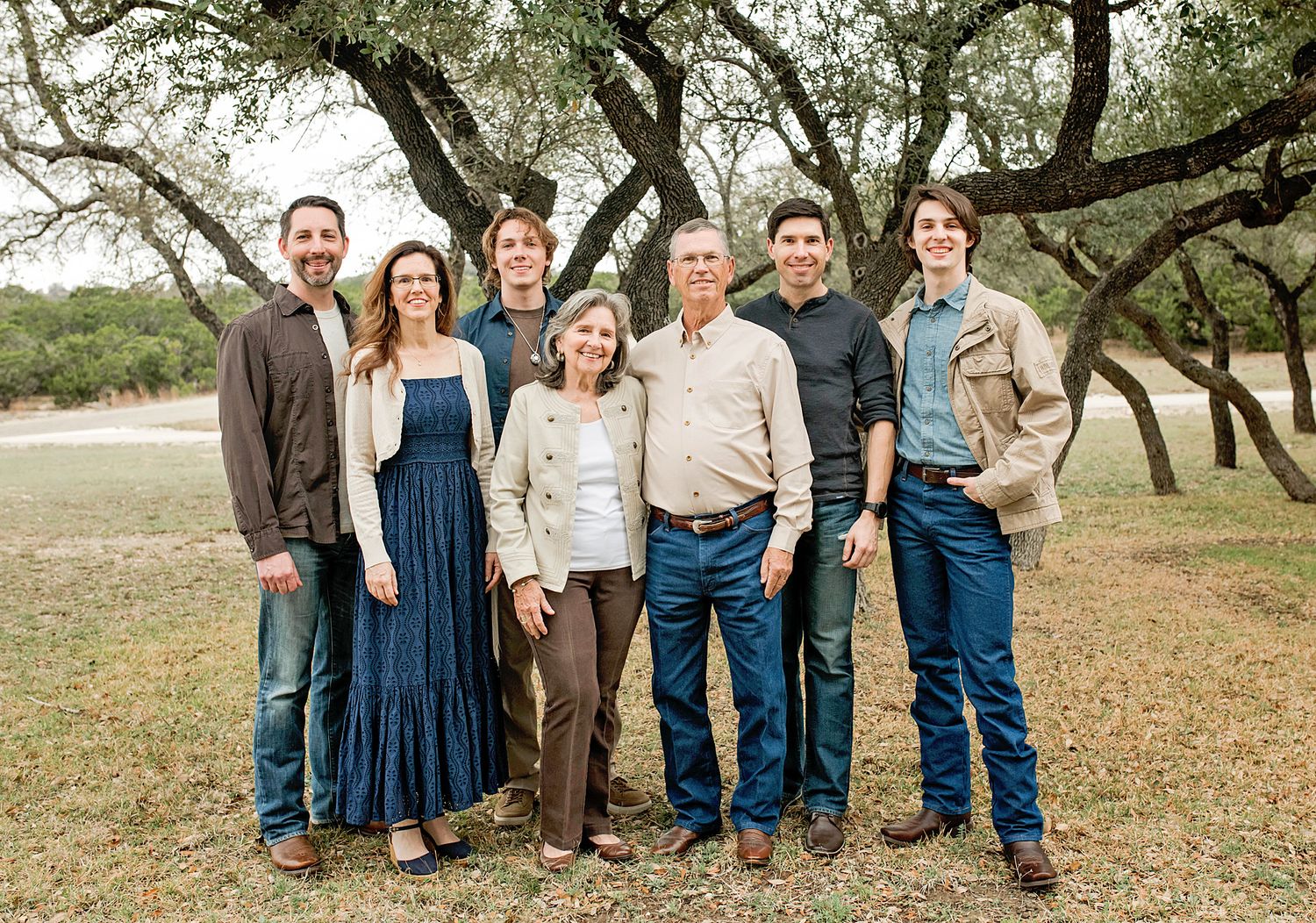 Family of seven adults smiling together in a wooded outdoor setting on a sunny day.