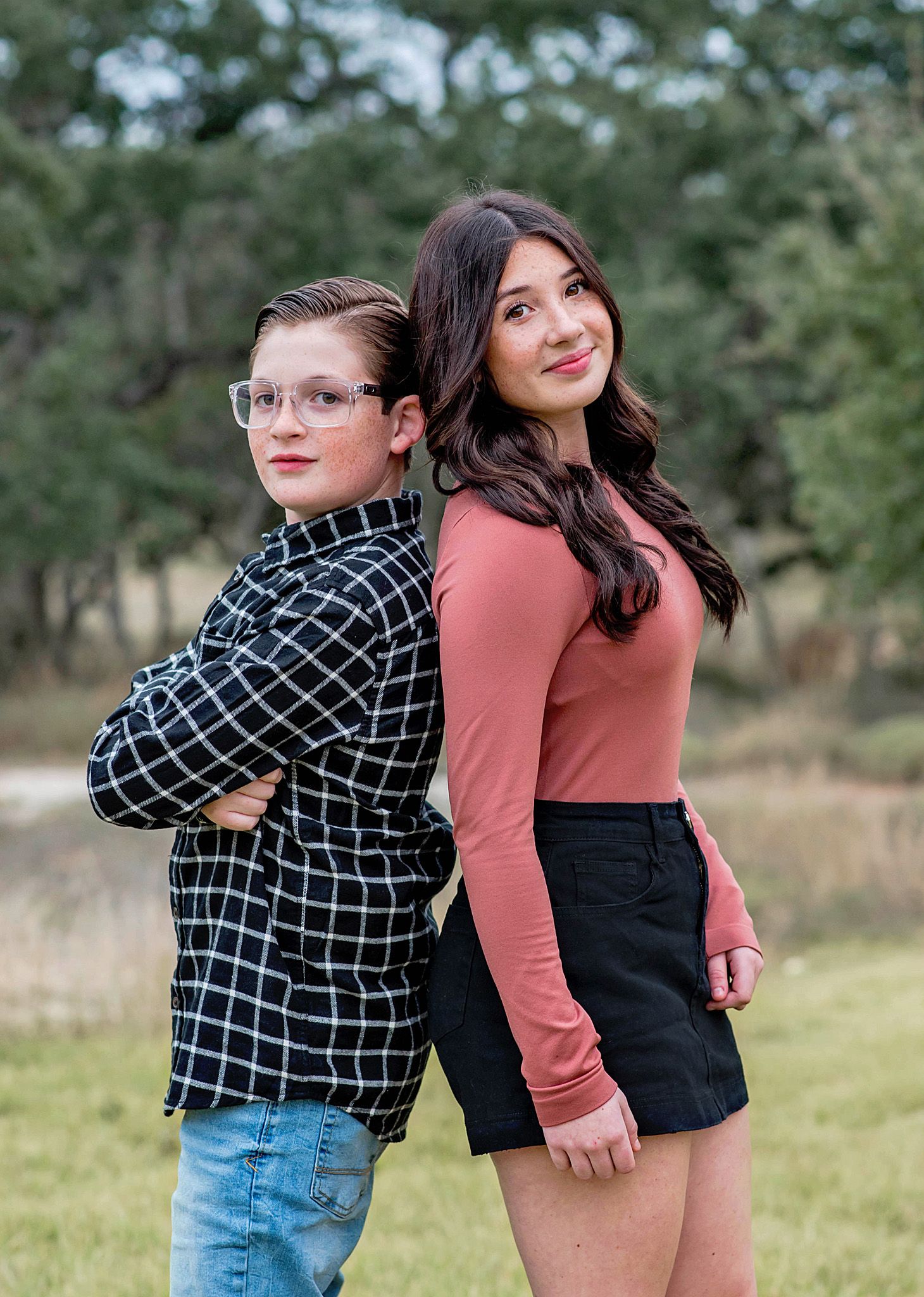 Two teenagers pose back-to-back outdoors in a casual fall portrait session in a natural setting.