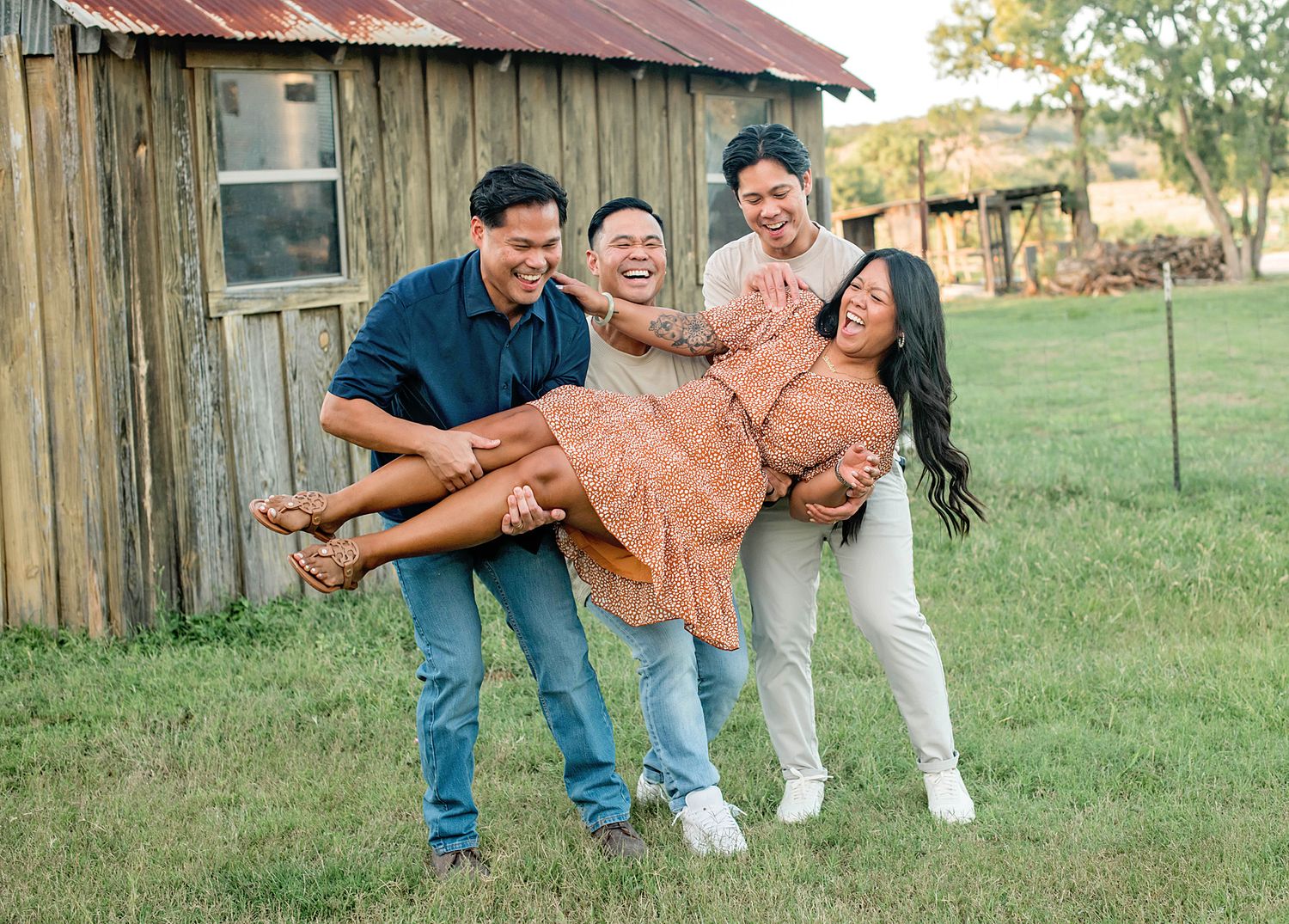 Four friends laugh outdoors near a rustic barn as two men playfully carry a smiling woman in an orange dress.