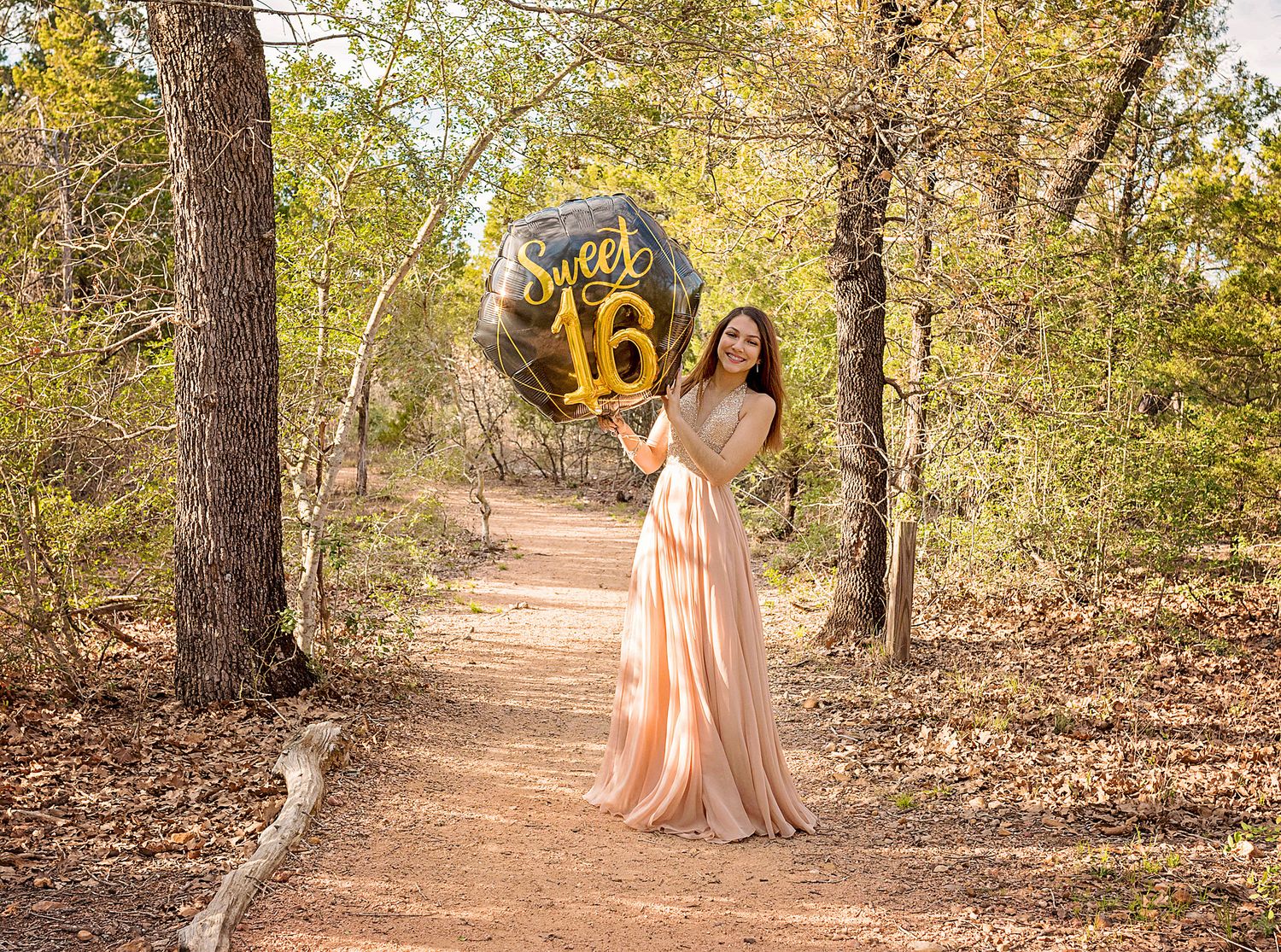 Teen girl in pink gown holding Sweet 16 gold balloon on a scenic forest path.