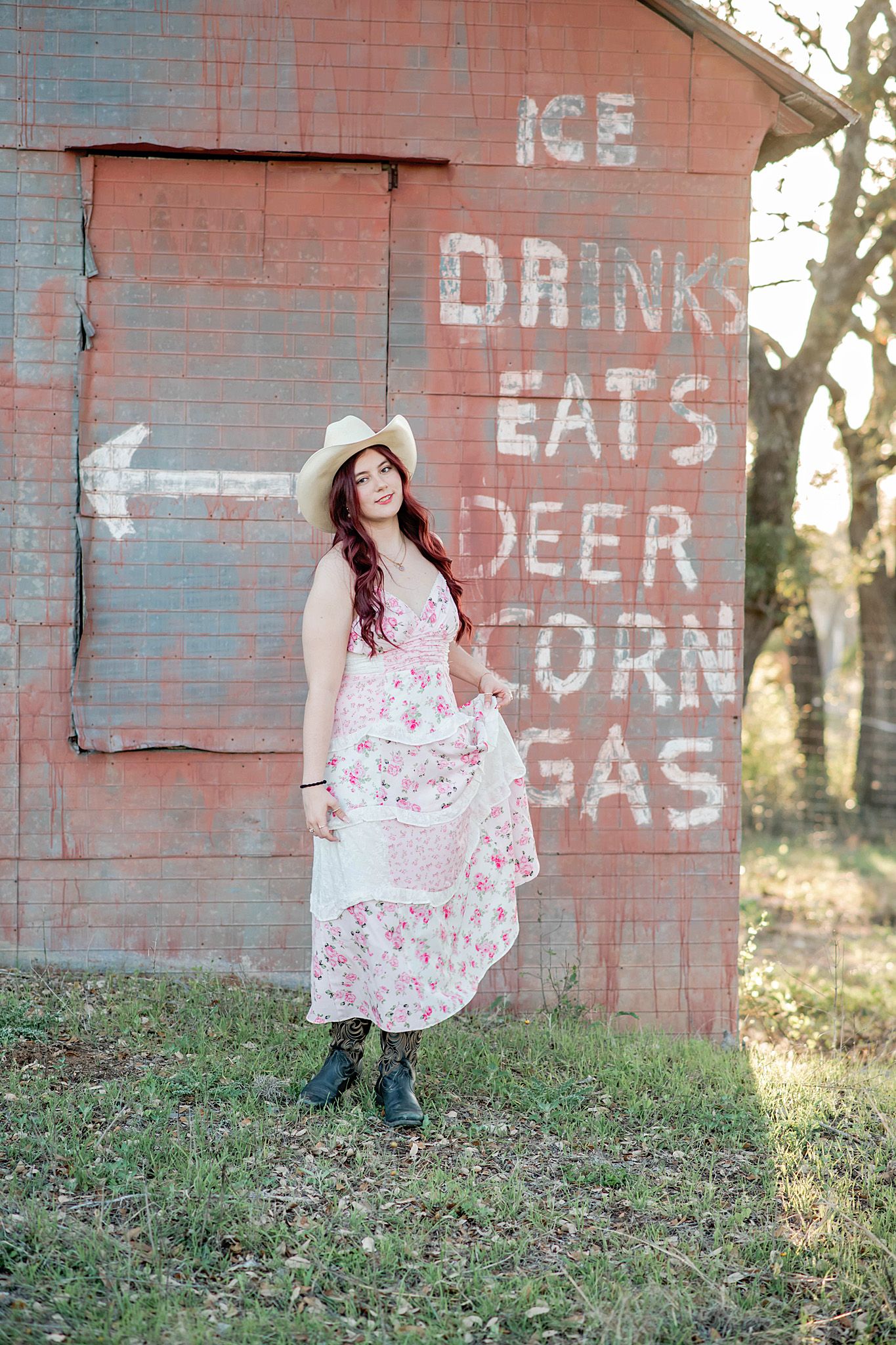 Woman in floral dress and cowboy hat poses by rustic red barn with painted signs for ice, drinks, eats, deer, corn, and gas.