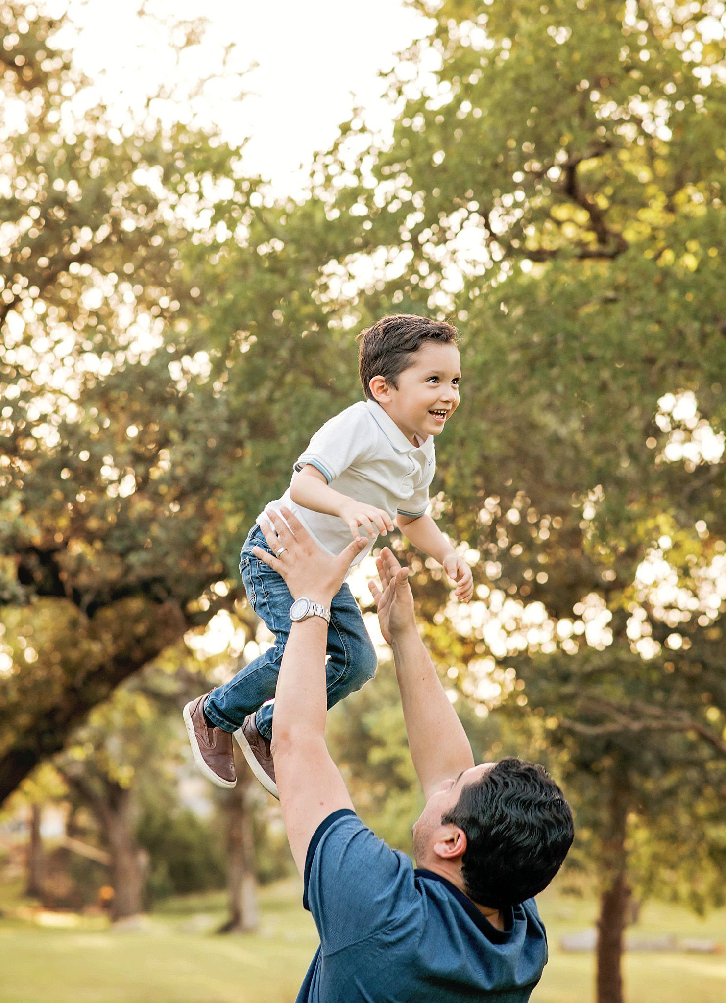 A parent lifts a young child high in the air during a playful moment in a sunlit park with trees in the background in Blanco, TX.