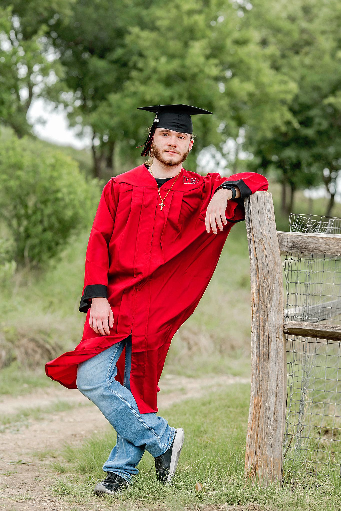 A person in a red graduation gown and cap poses casually against a wooden fence post in an outdoor rural setting.