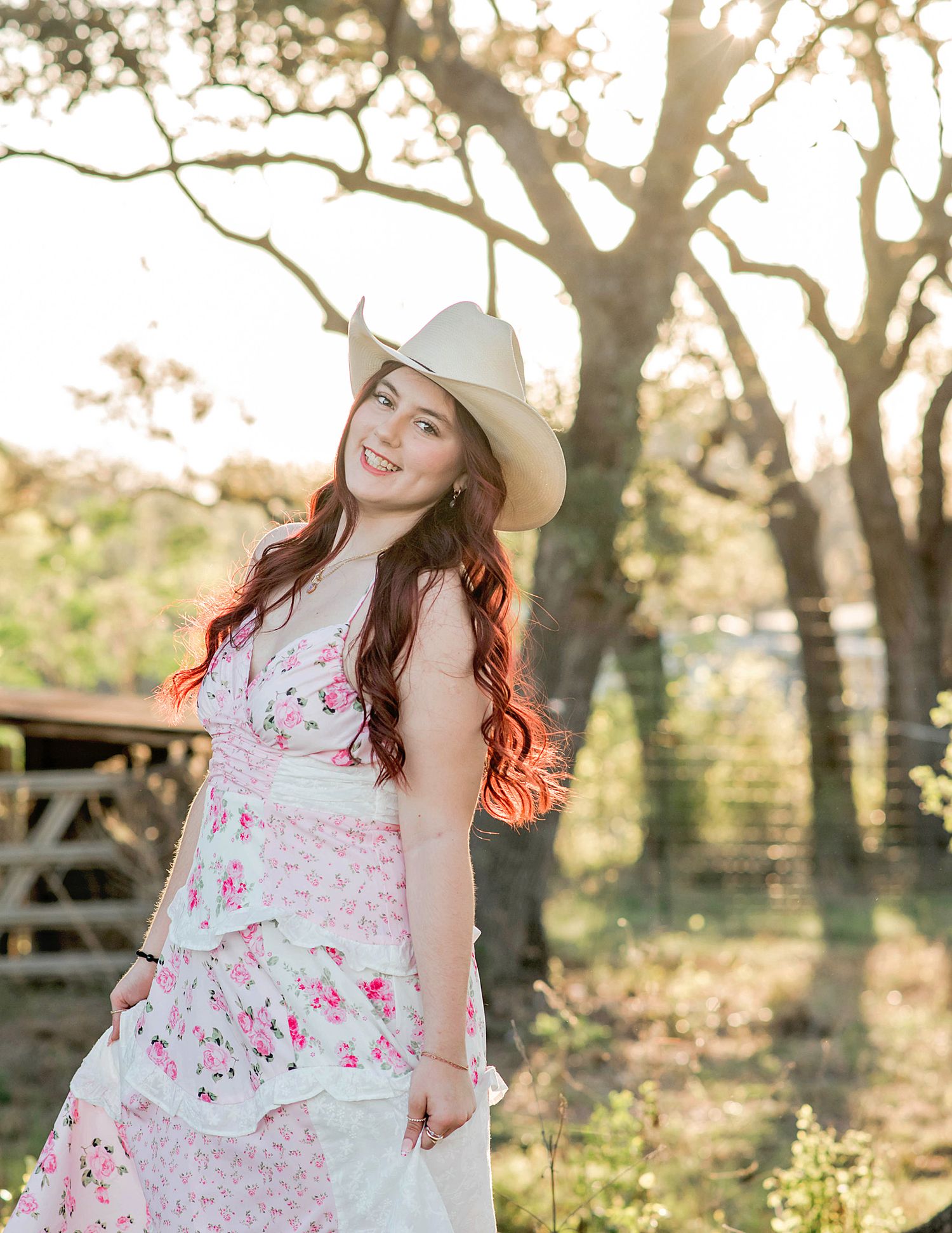 Young woman in floral dress and white cowboy hat smiling outdoors in golden sunlight near trees.