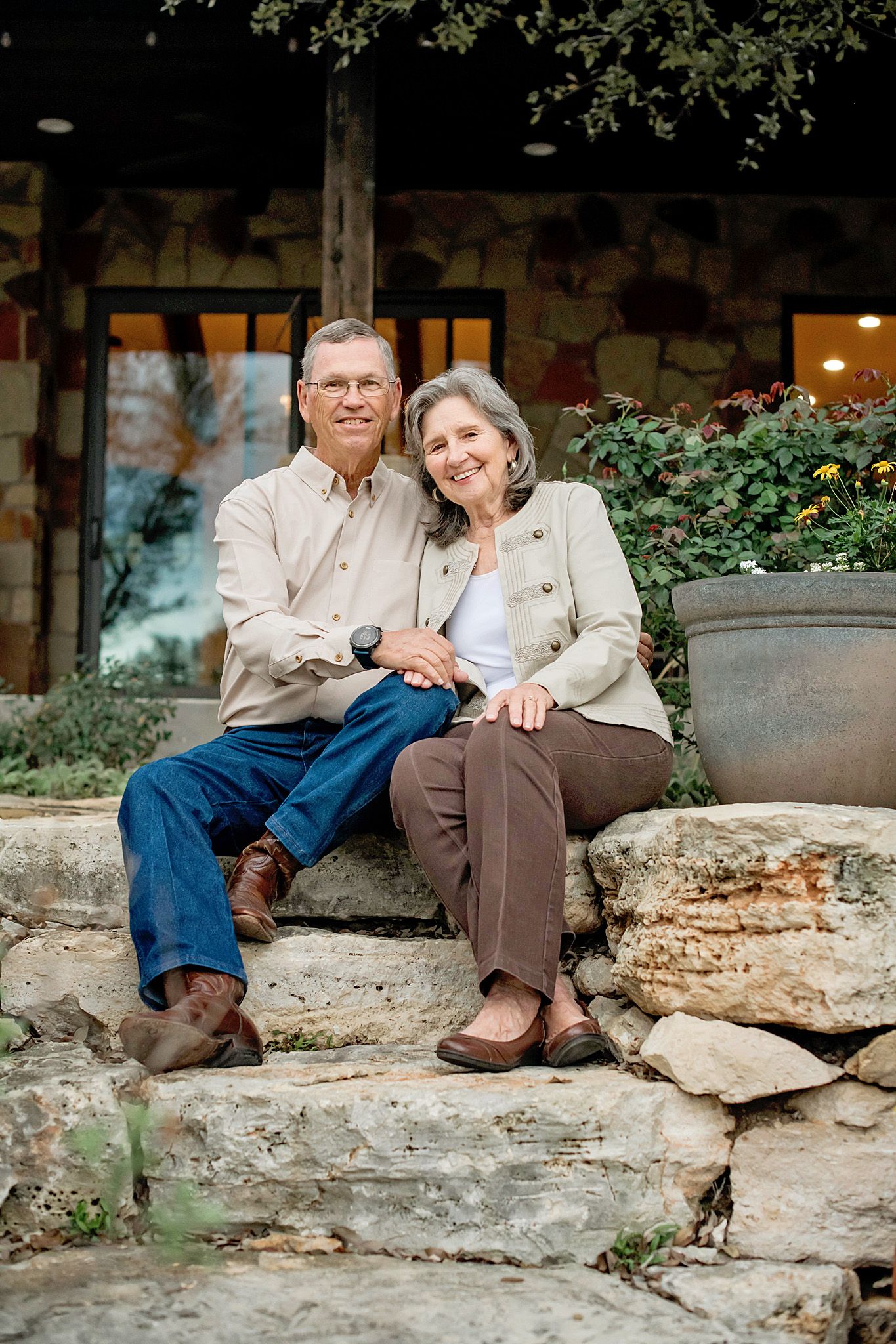Smiling senior couple sitting together on stone steps outside a rustic home surrounded by greenery.