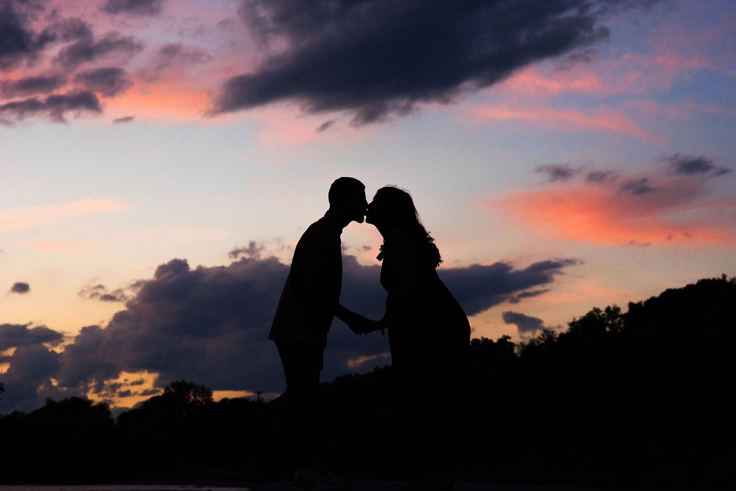 Silhouettes of a couple sharing an intimate moment against a vibrant sunset sky with dark clouds in Blanco, TX.