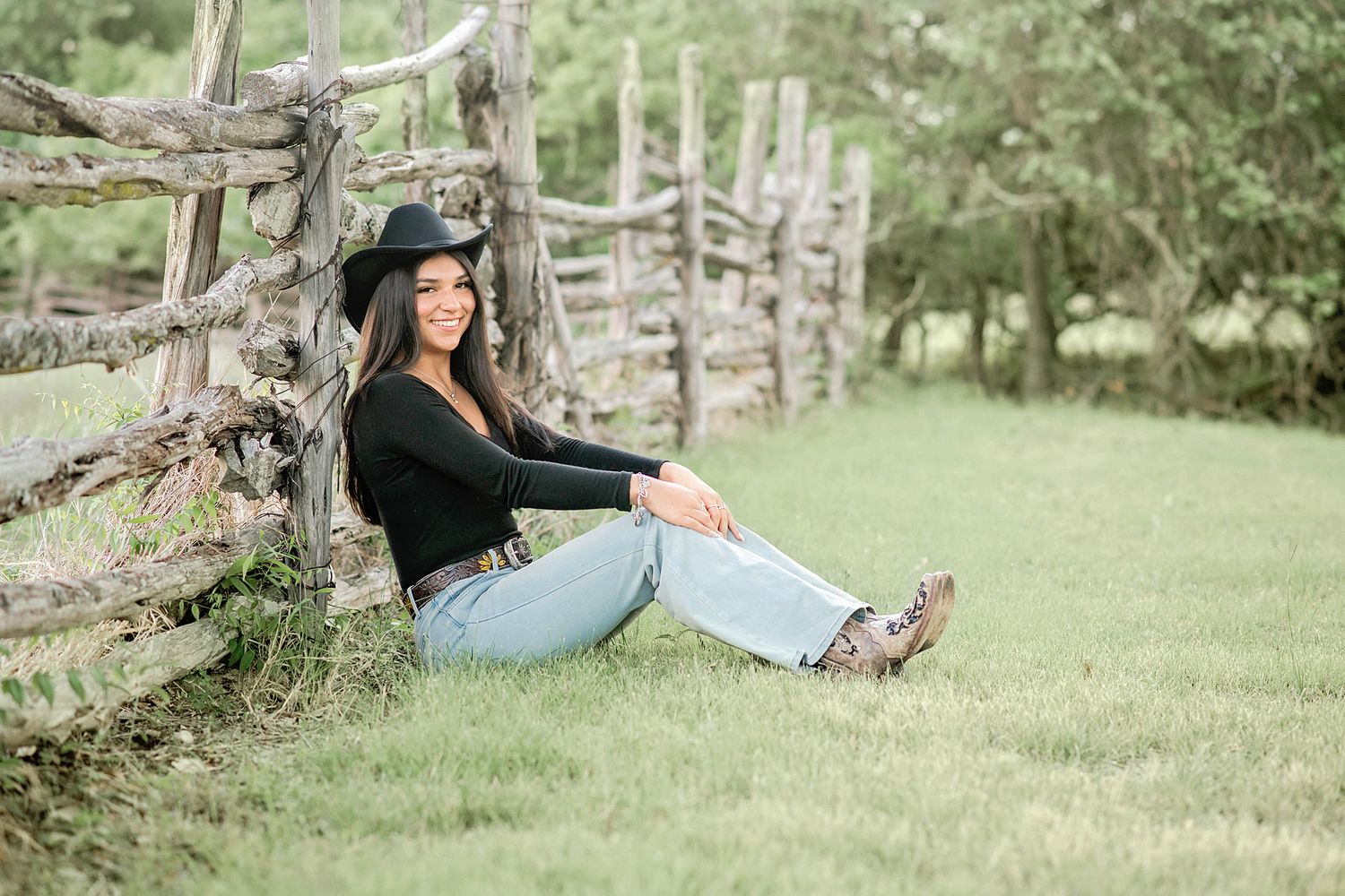 Young woman in black hat and jeans sits smiling against rustic wooden fence on green grass outdoors.