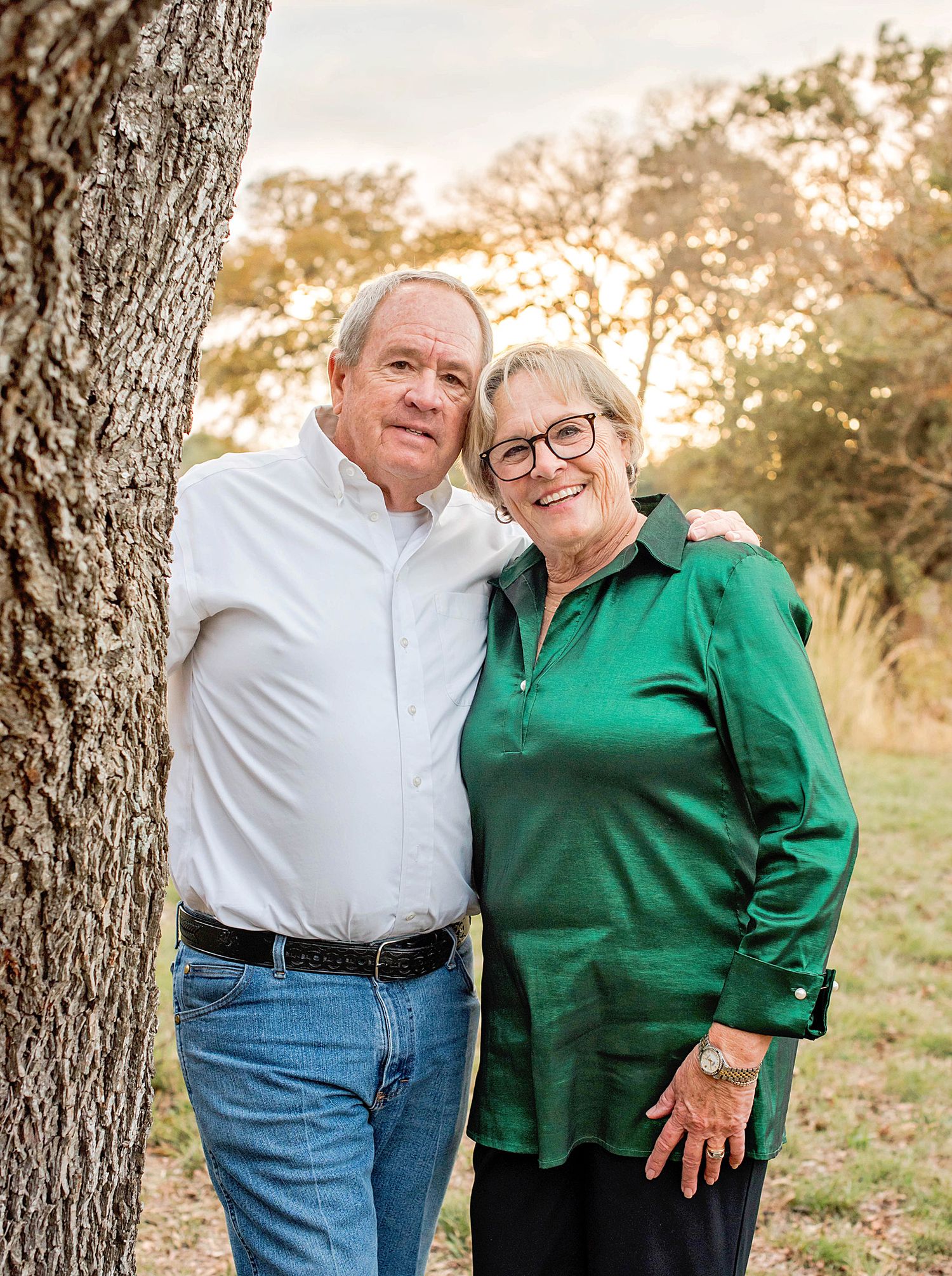 Loving senior couple in white shirt and green top embrace while standing next to a tree trunk at sunset.