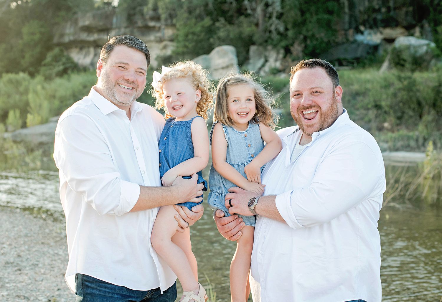 Two men in white shirts smiling outdoors, each holding a young girl in blue dresses near a creek.
