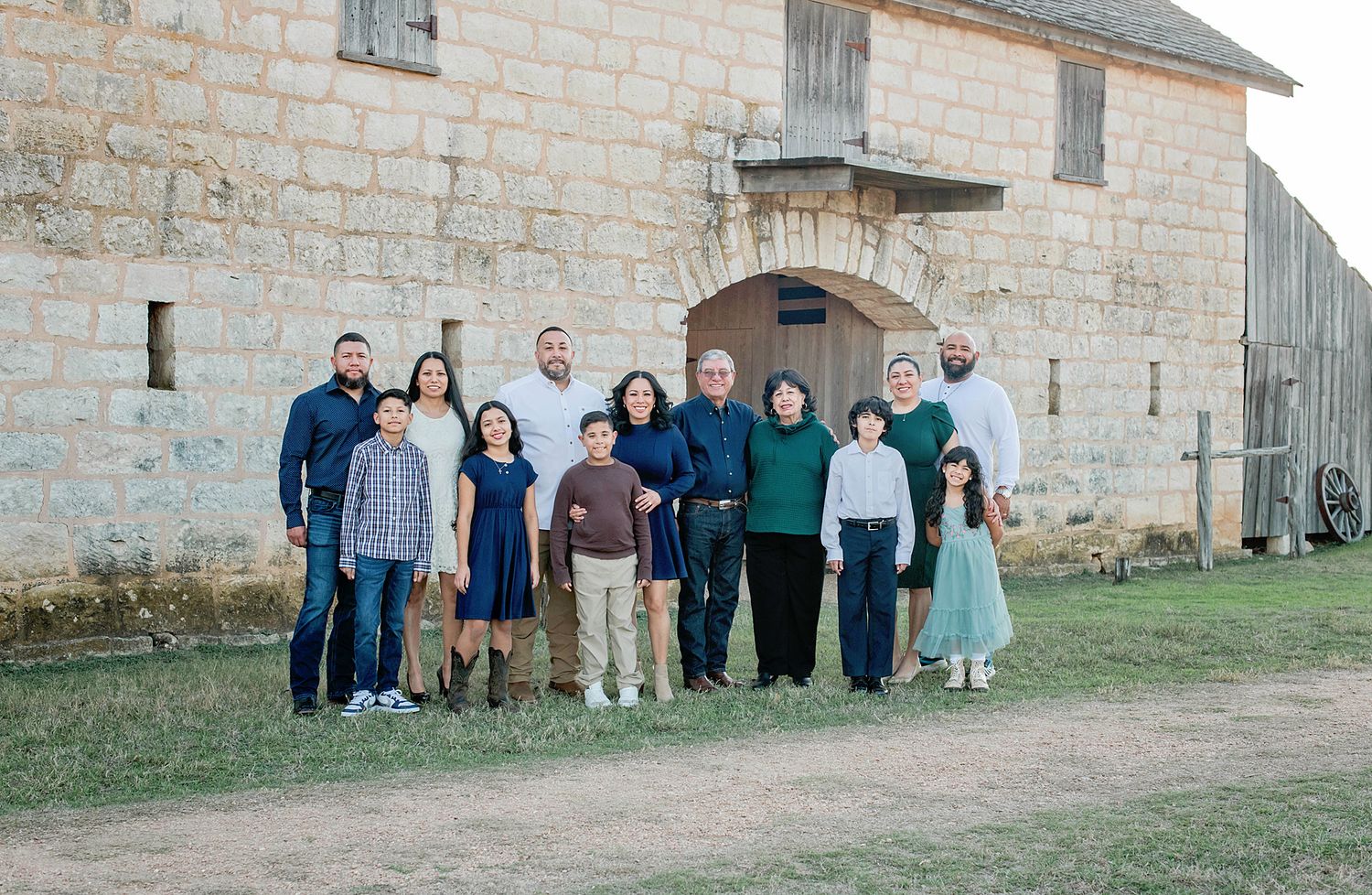 Large family group portrait posed outdoors near a rustic stone barn building on a sunny day.