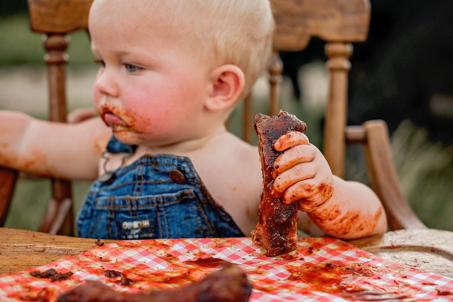 A toddler in overalls enjoying a messy barbecue meal with sauce-covered hands and face.
