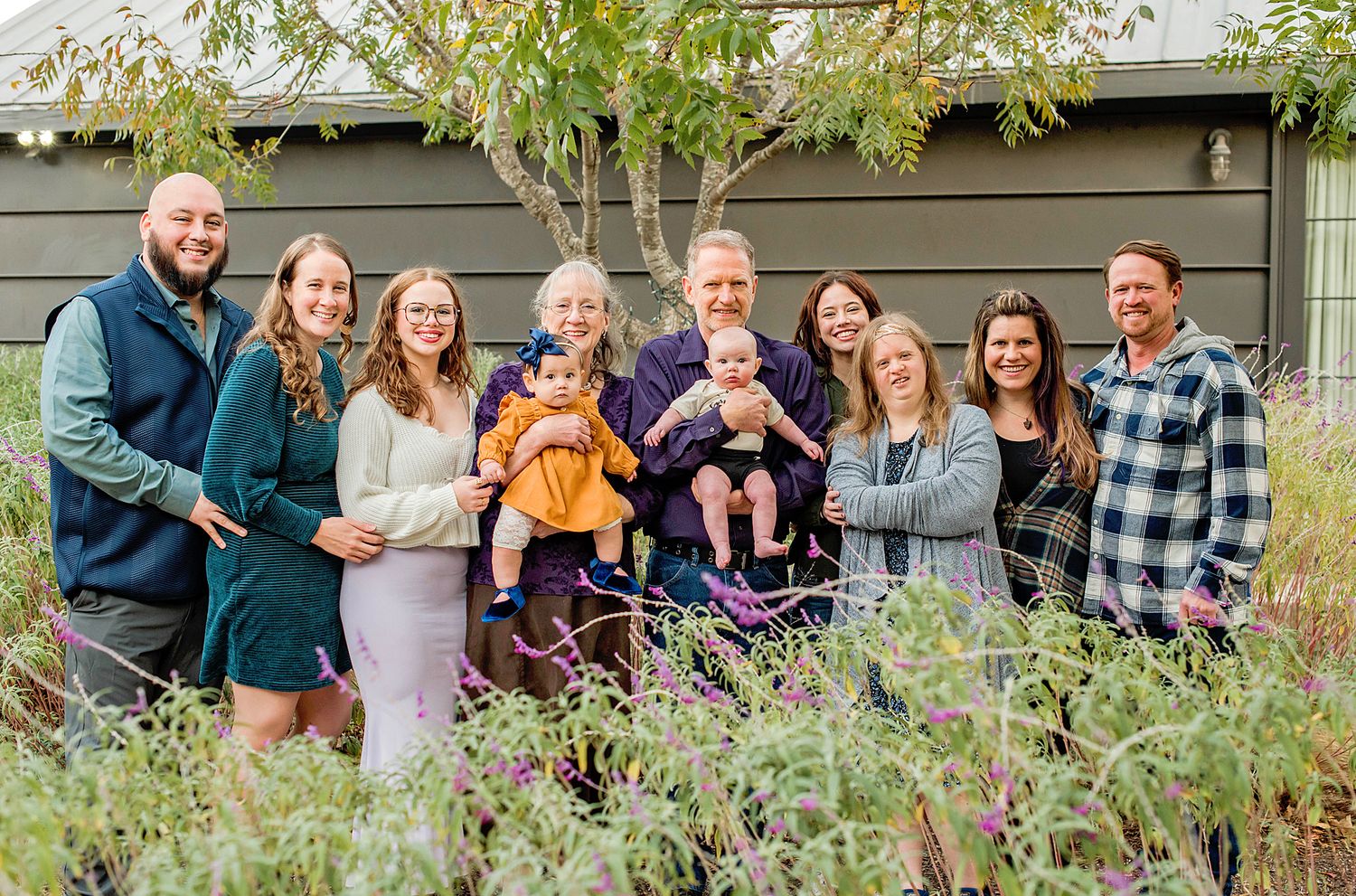 A large multigenerational family poses together outdoors in a lush garden, including two babies and smiling adults.