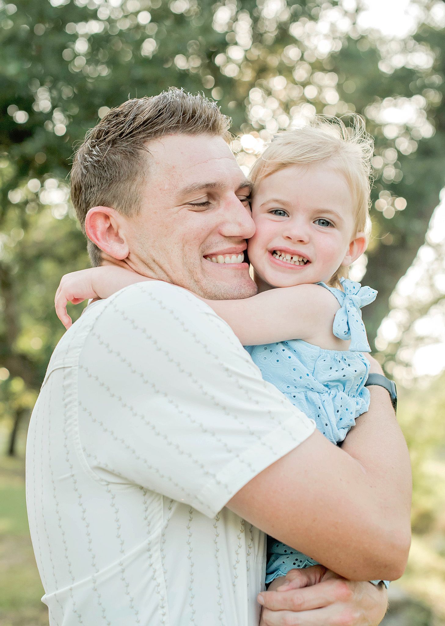 A smiling man hugs a happy young blonde girl in a blue dress outdoors in a sunny park setting.