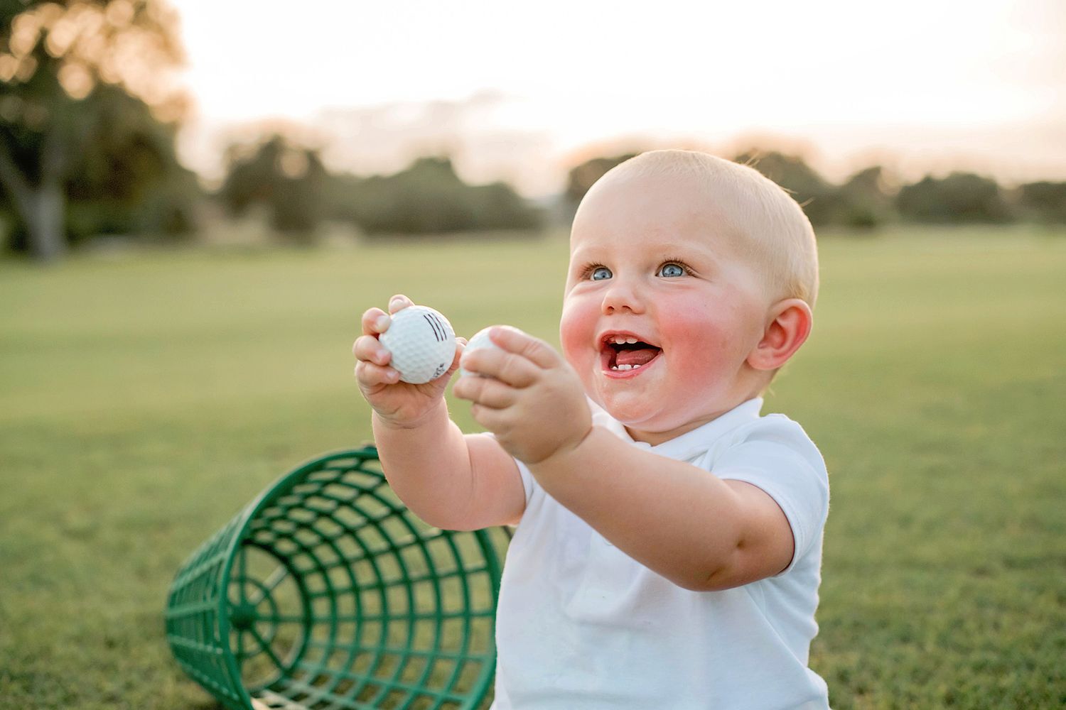 Child wearing white shirt laughs joyfully while holding a baseball on sunny grass field.