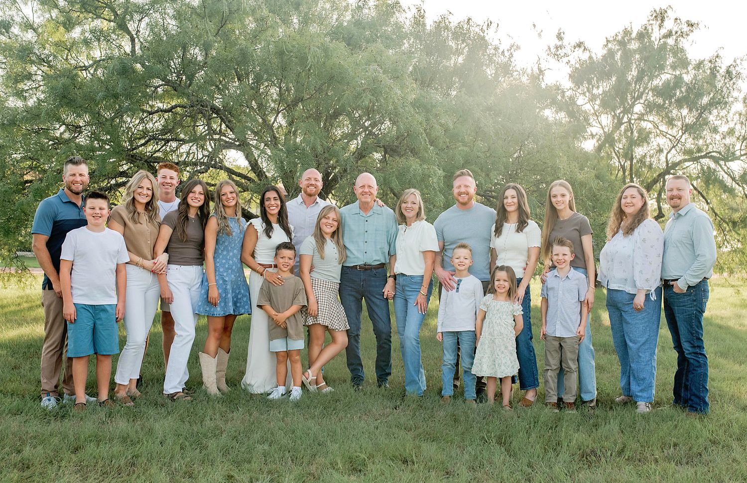 Large family group portrait outdoors on green grass with trees, dressed in coordinated casual outfits.