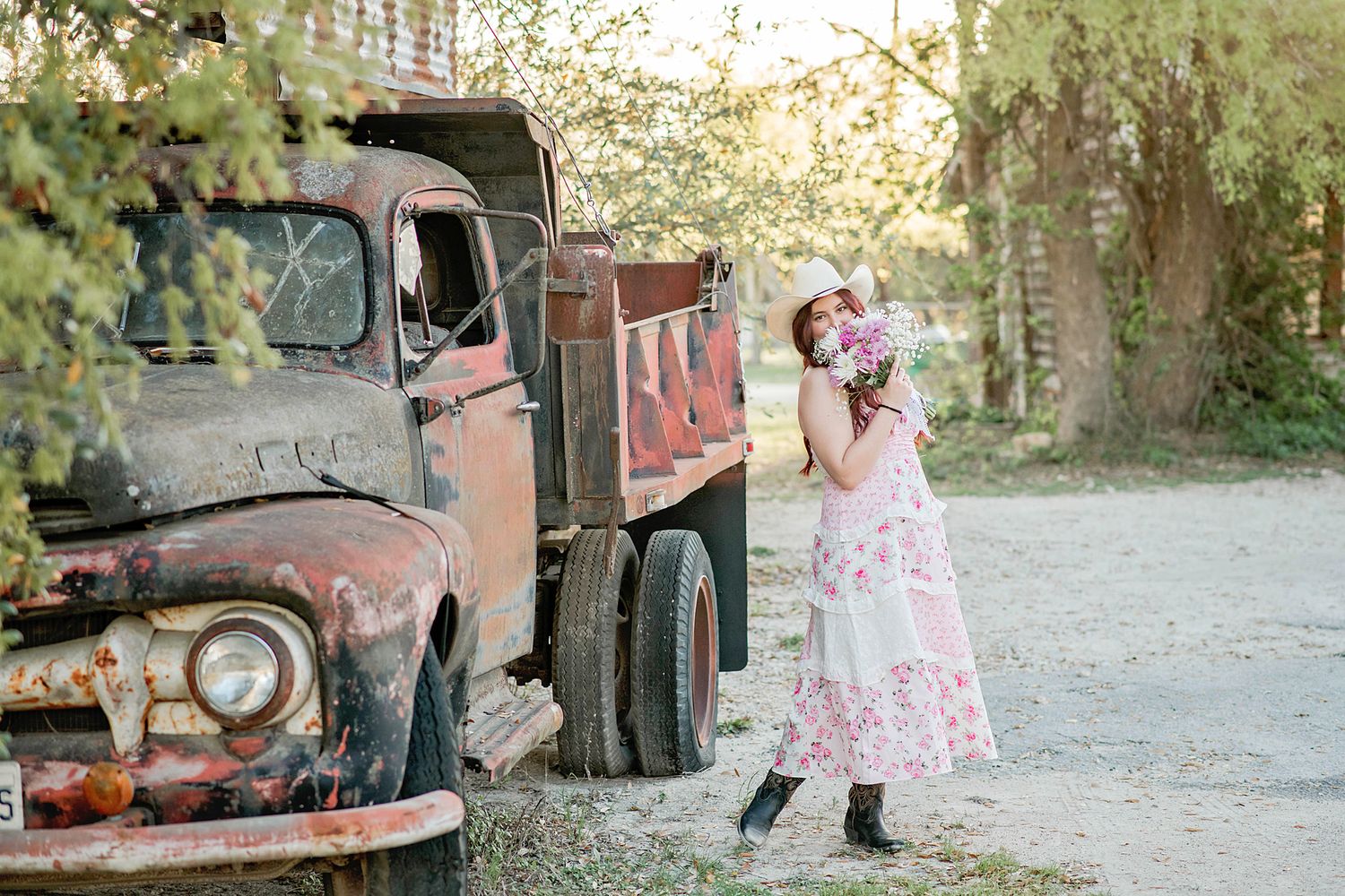 Woman in floral dress holding bouquet beside rusty vintage truck outdoors.