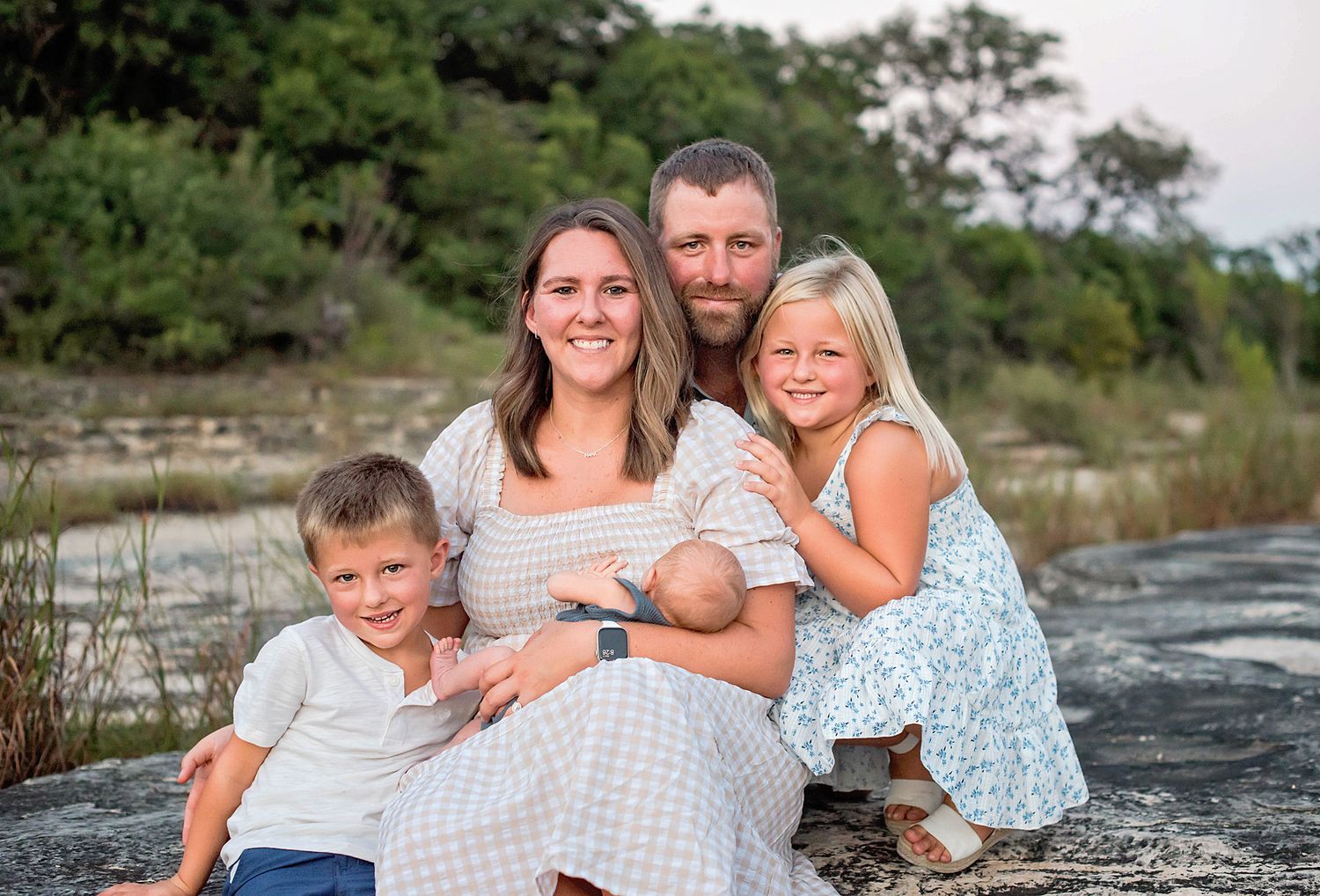 Family relaxing on rocky shore beside water, dressed in coordinating blue and white outfits.
