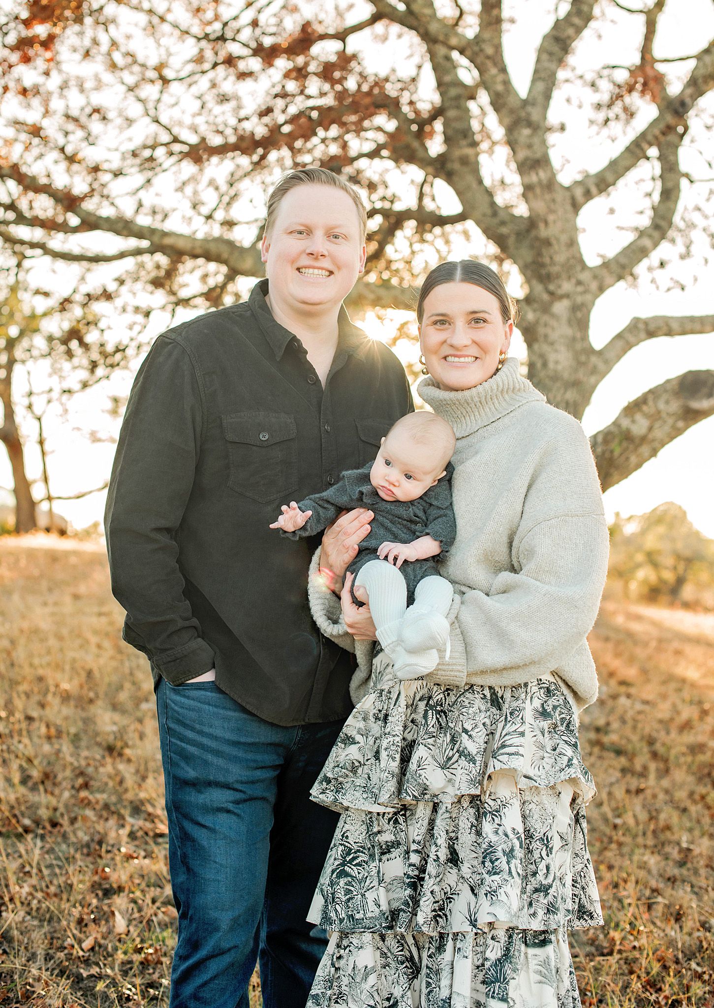 Family portrait of couple holding infant baby outdoors under oak tree in golden sunset light.