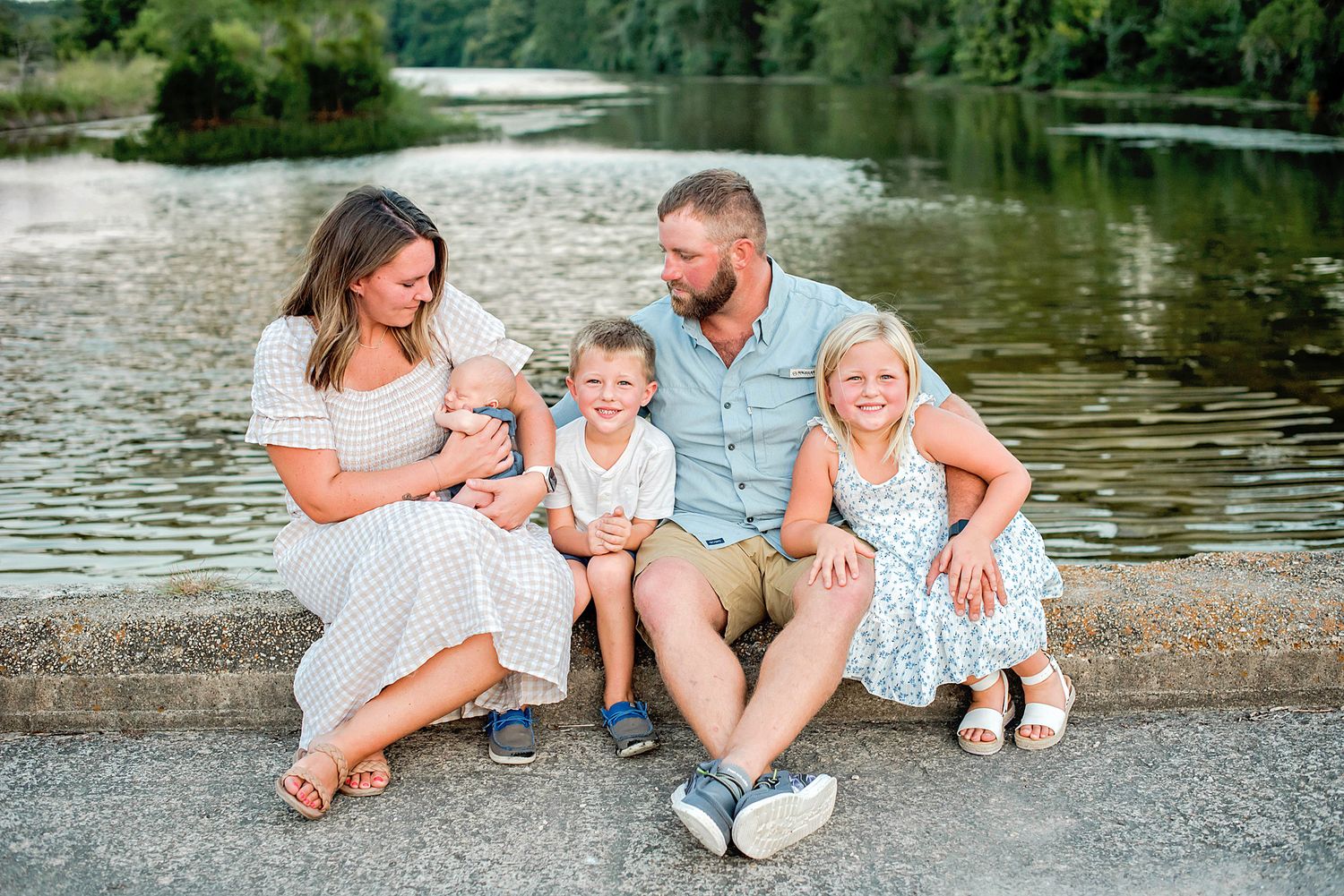 A family sits together on a stone wall by a serene lake during a summer afternoon.