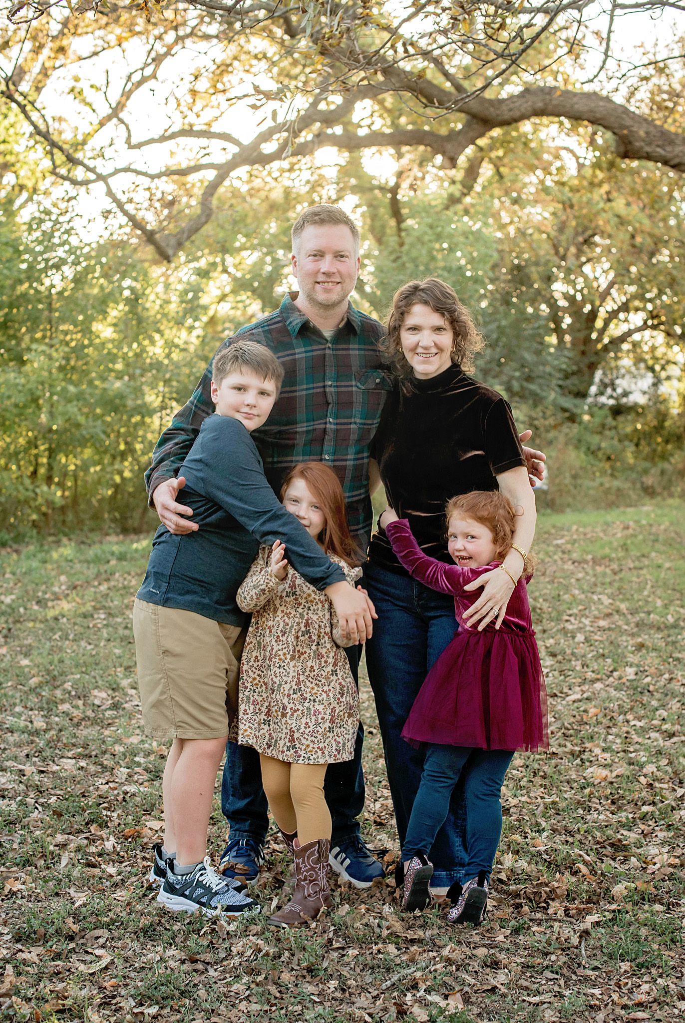 Family of five posing outdoors in fall foliage, parents with three young children smiling under a large tree.