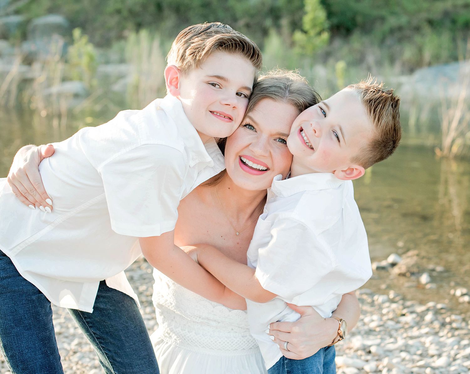 A smiling mother hugs her two young sons in white shirts outdoors near a rocky riverbank.