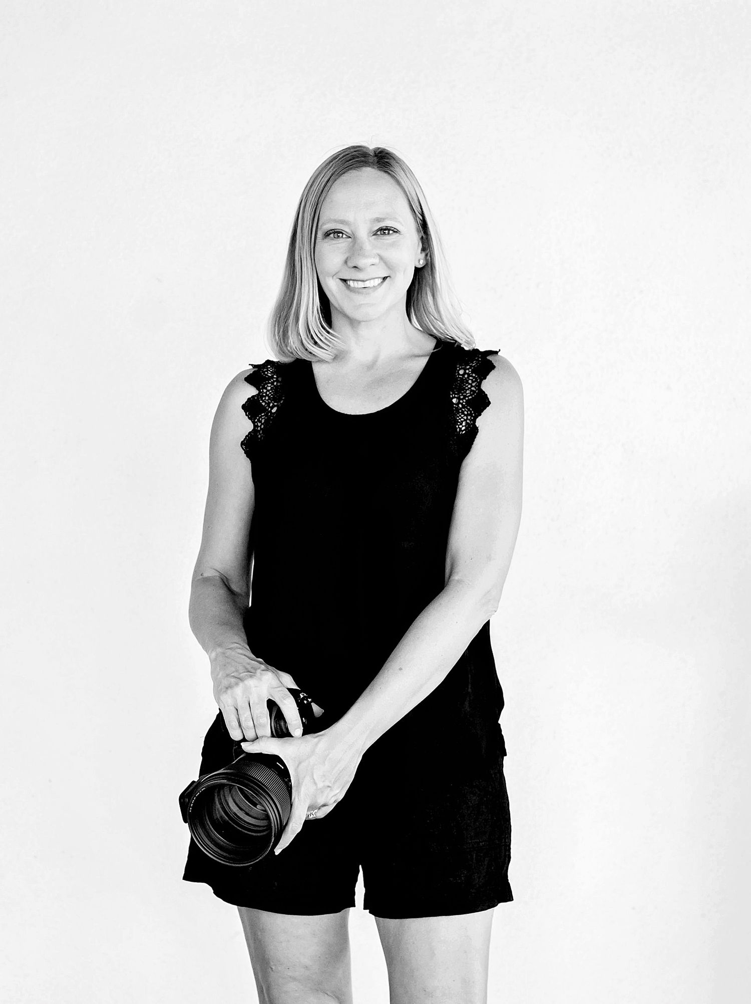 A photographer in black attire holds a professional camera against a white background.