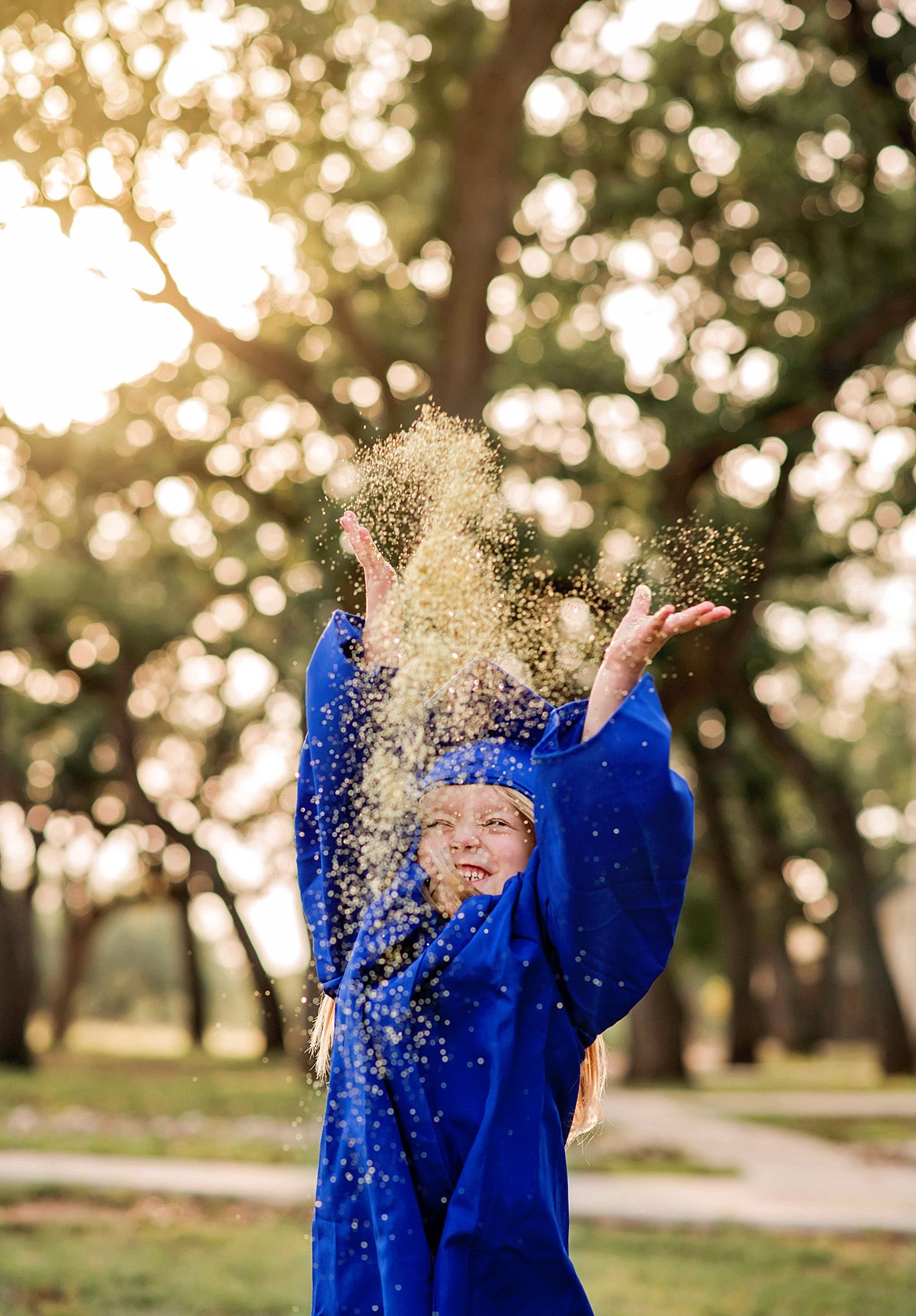 Graduate in blue gown tosses glitter in sunlit park, celebrating academic achievement outdoors.