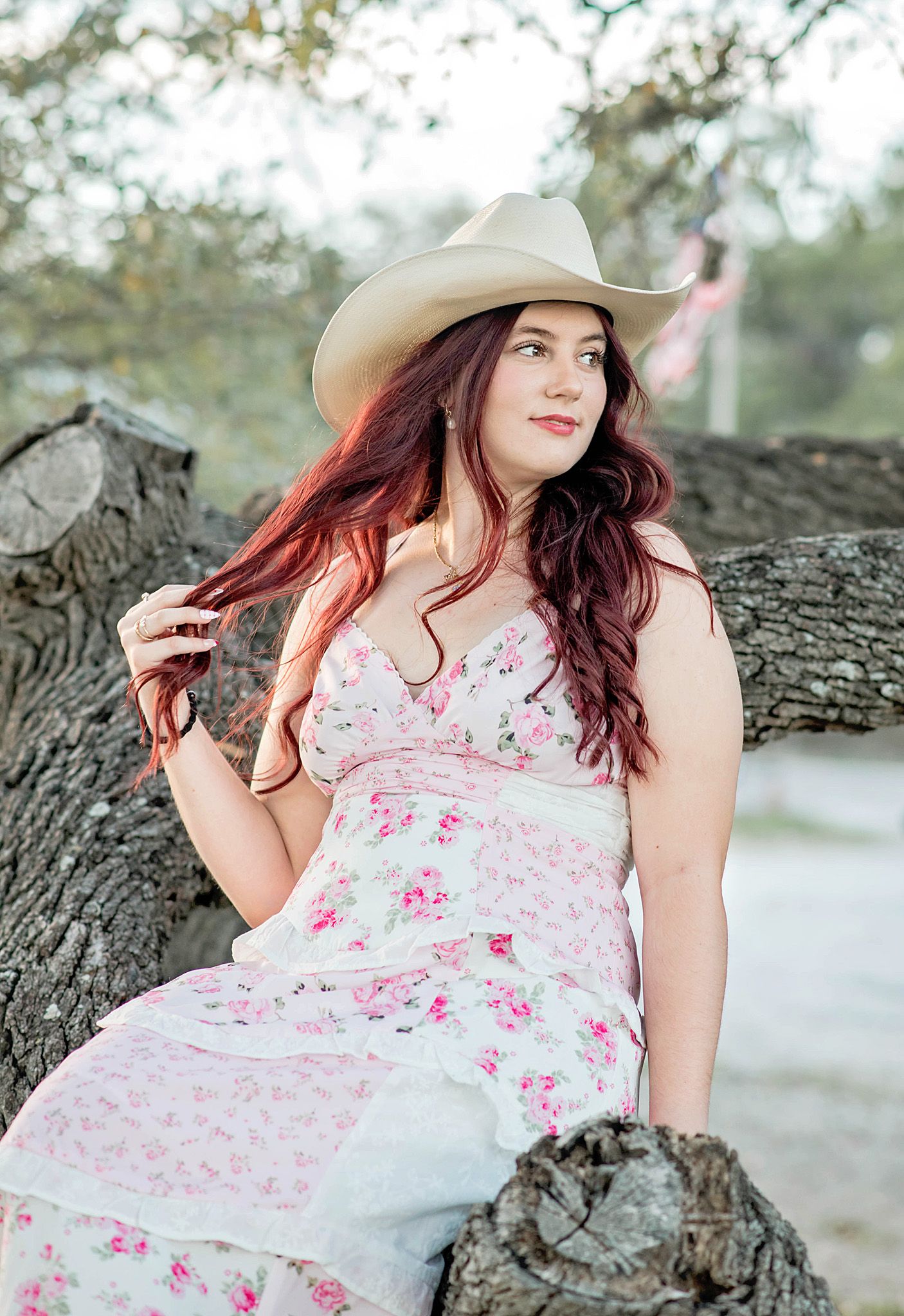 Young woman with red hair in floral dress and cowboy hat sitting on a tree trunk outdoors.