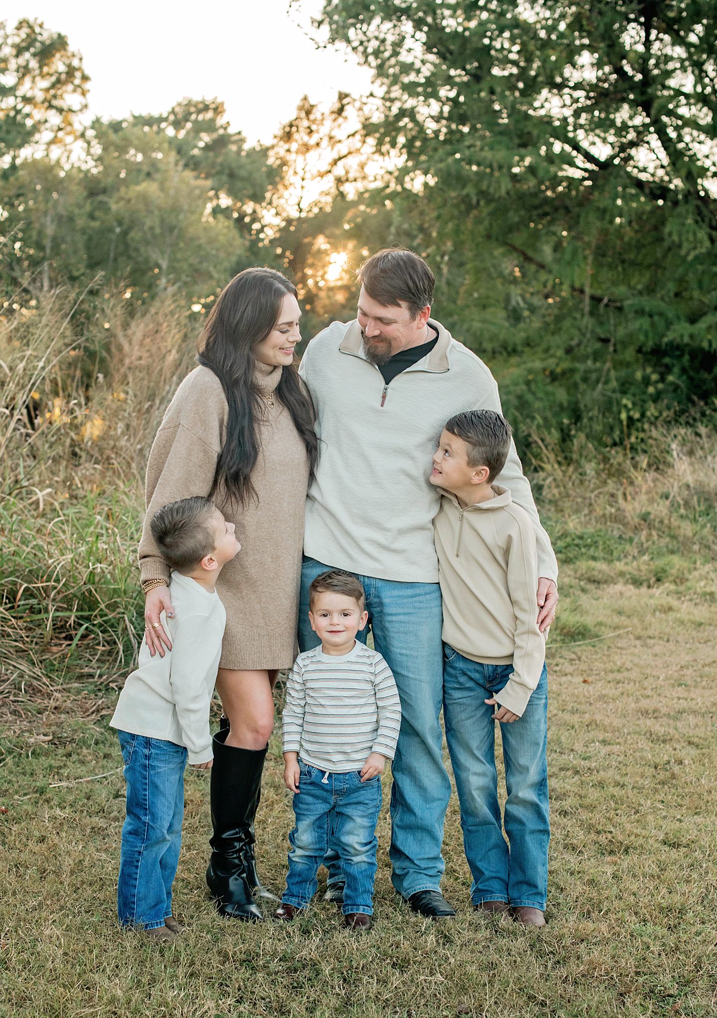 A family in casual attire stands together on grass during golden hour in a natural outdoor setting.