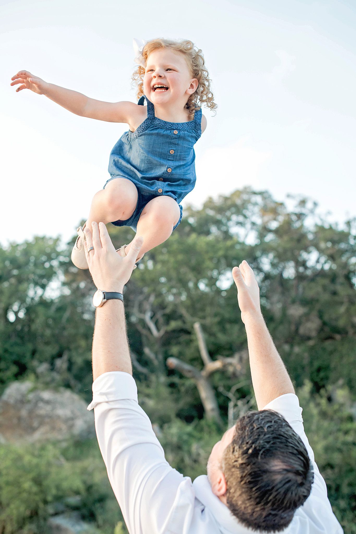 Father joyfully tosses laughing toddler girl in blue romper into the air outdoors with trees in background.