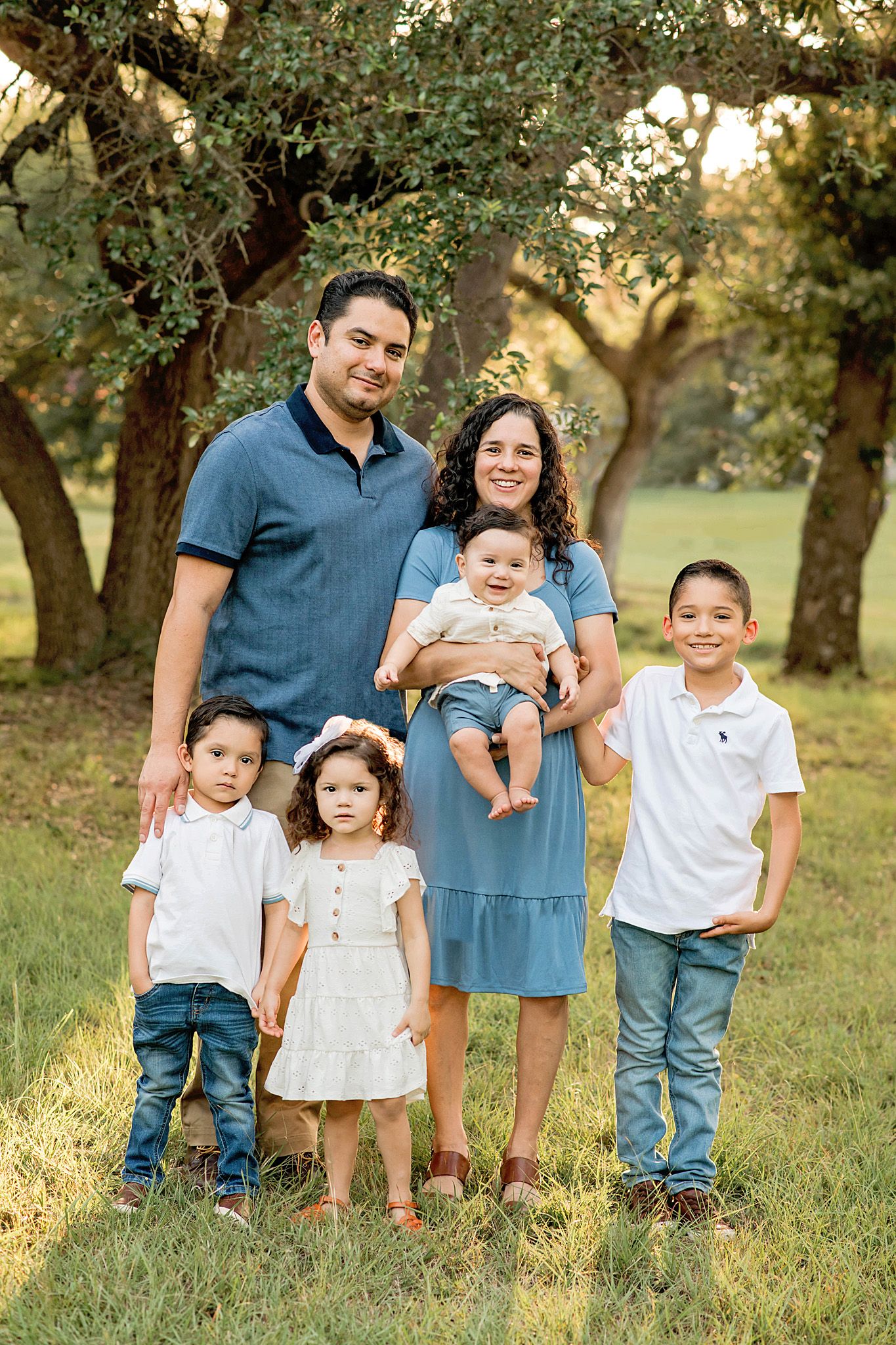 A family poses together outdoors in a grassy field with olive trees during golden hour lighting in Blanco, TX.