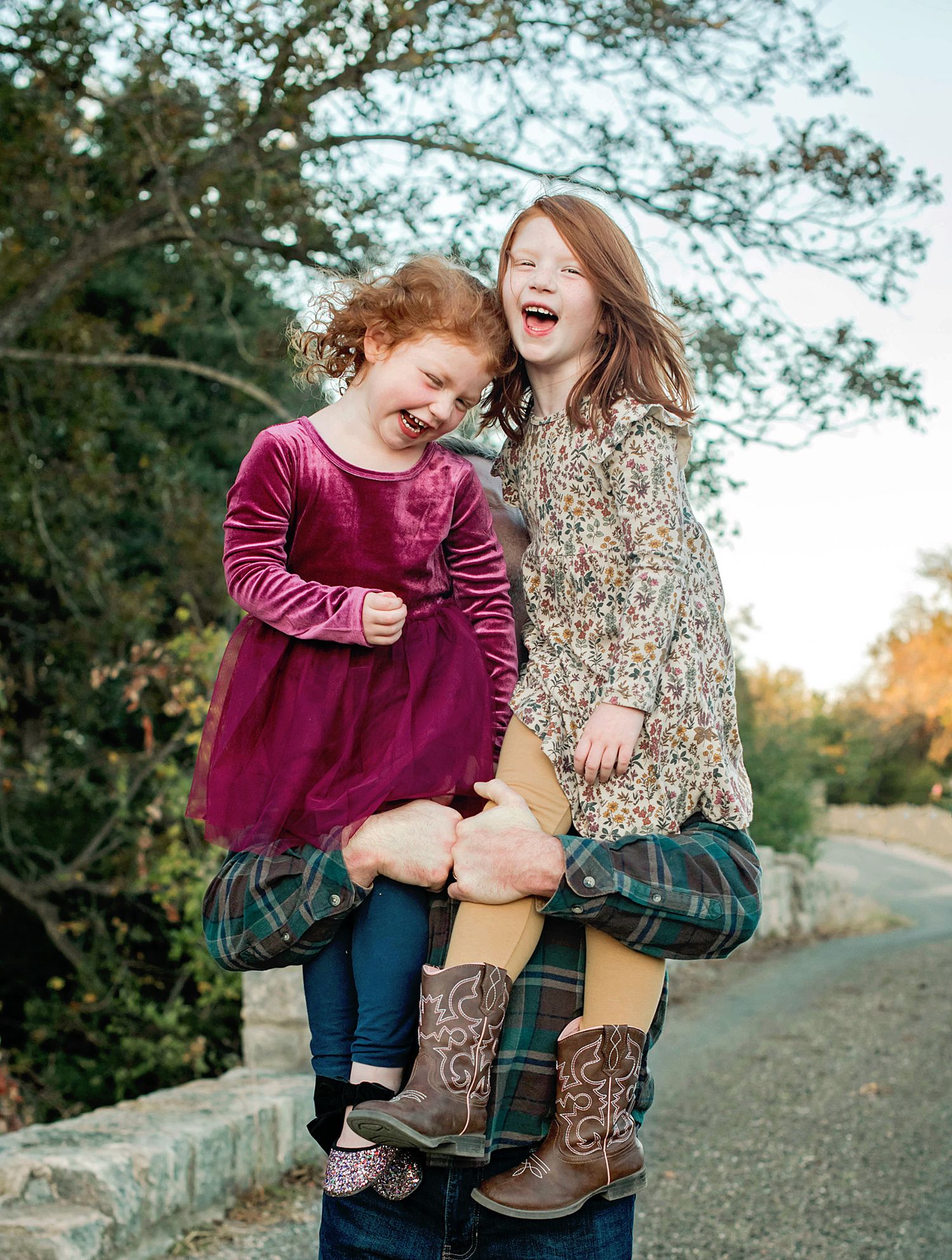 Two laughing girls in dresses and cowboy boots pose outdoors, one held up joyfully, surrounded by autumn trees.