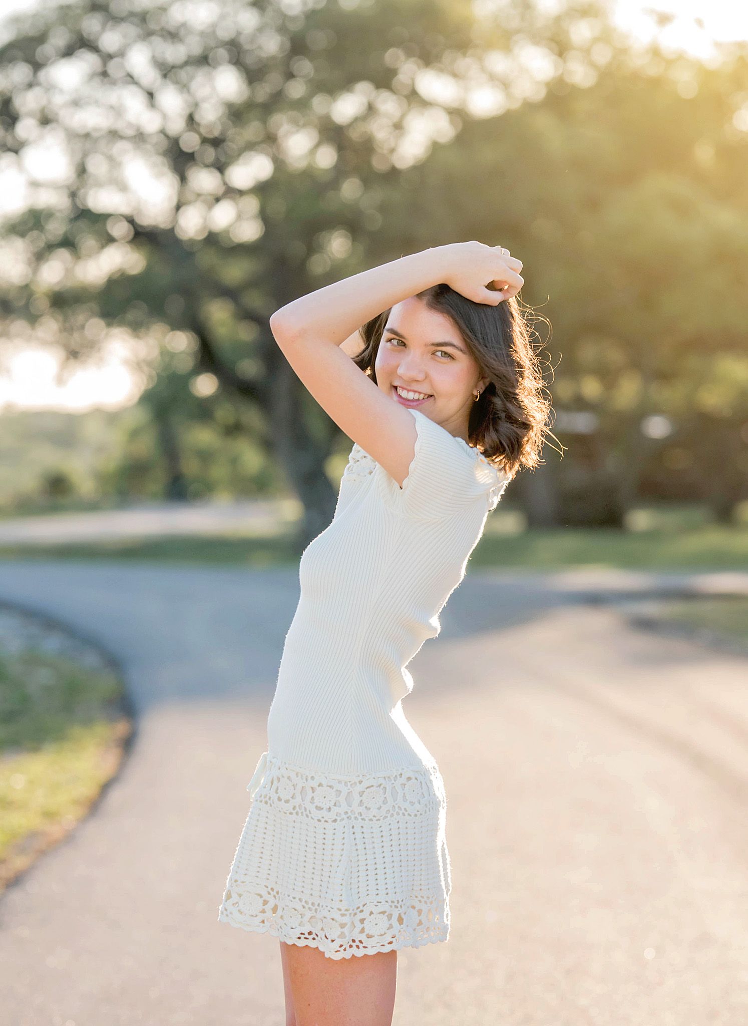 Young woman in white lace mini dress poses on a sunlit road with golden hour light and trees in background.
