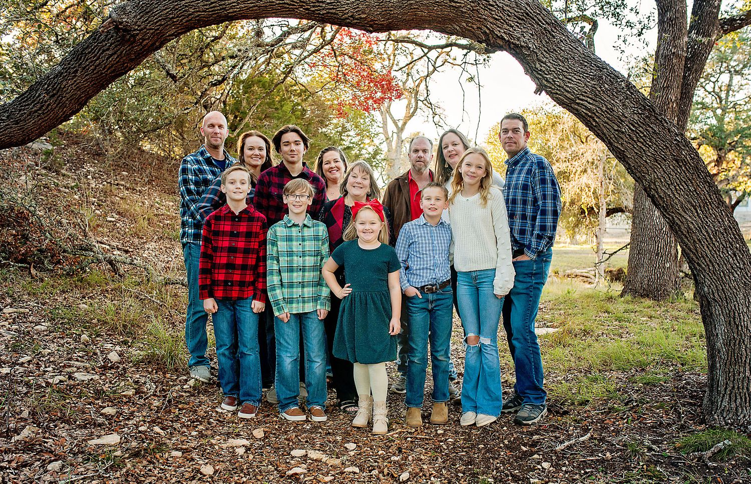 Large family of twelve smiling under a curved tree branch in fall foliage.