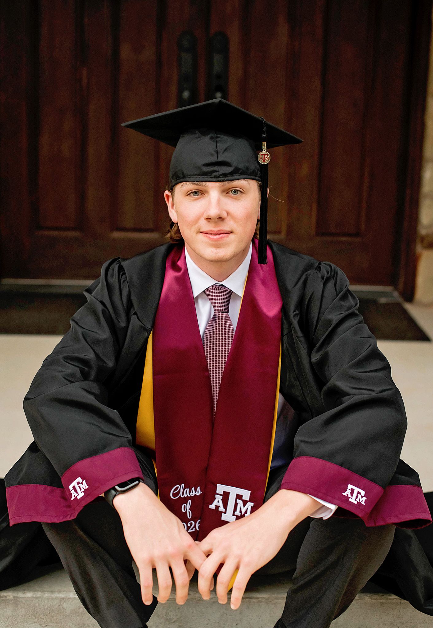 Texas A&M University graduate poses in cap and gown with maroon stole reading Class of 2026 on wooden steps.