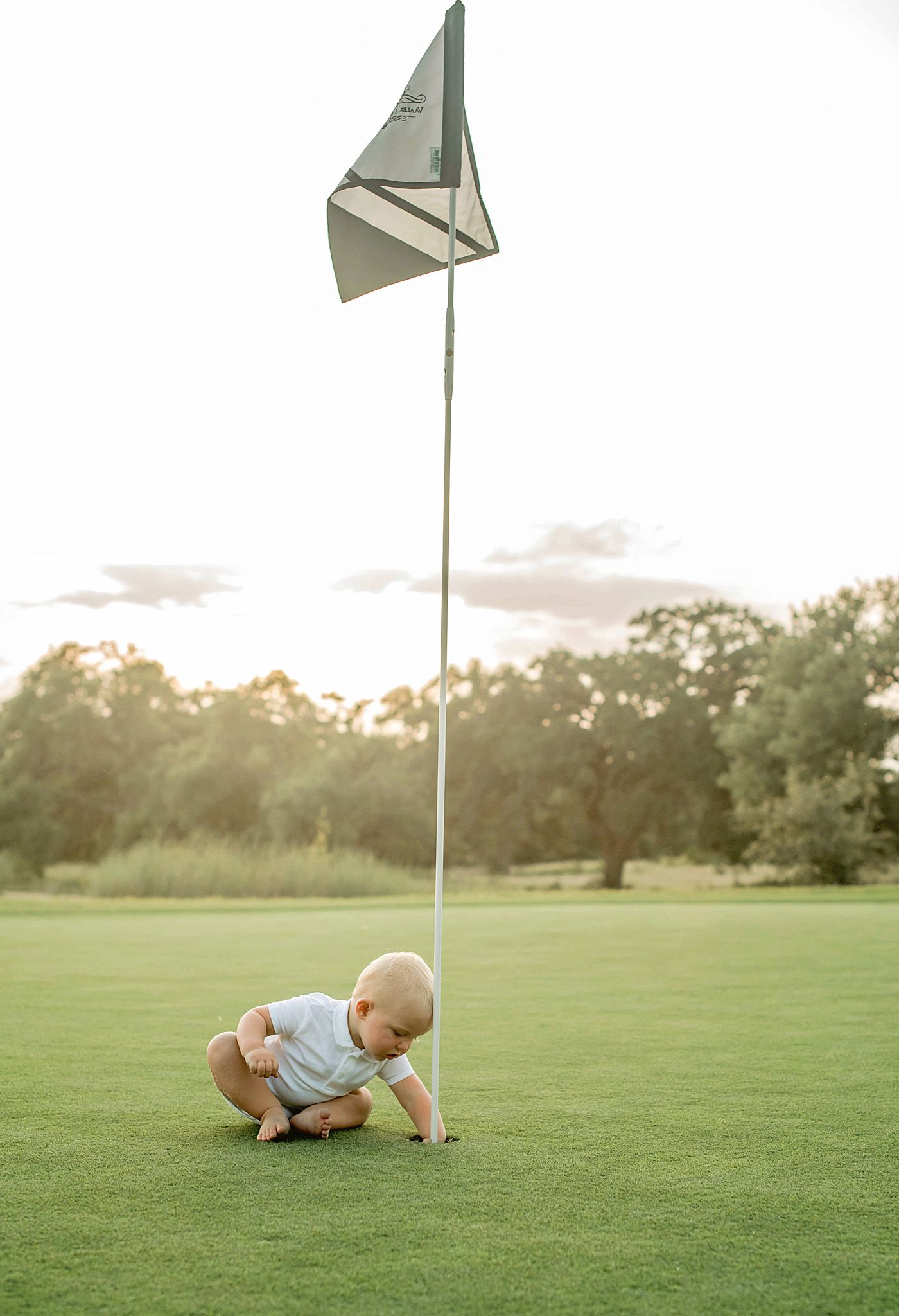 Baby in light-colored outfit crawls and plays on a green golf course during golden hour sunset.