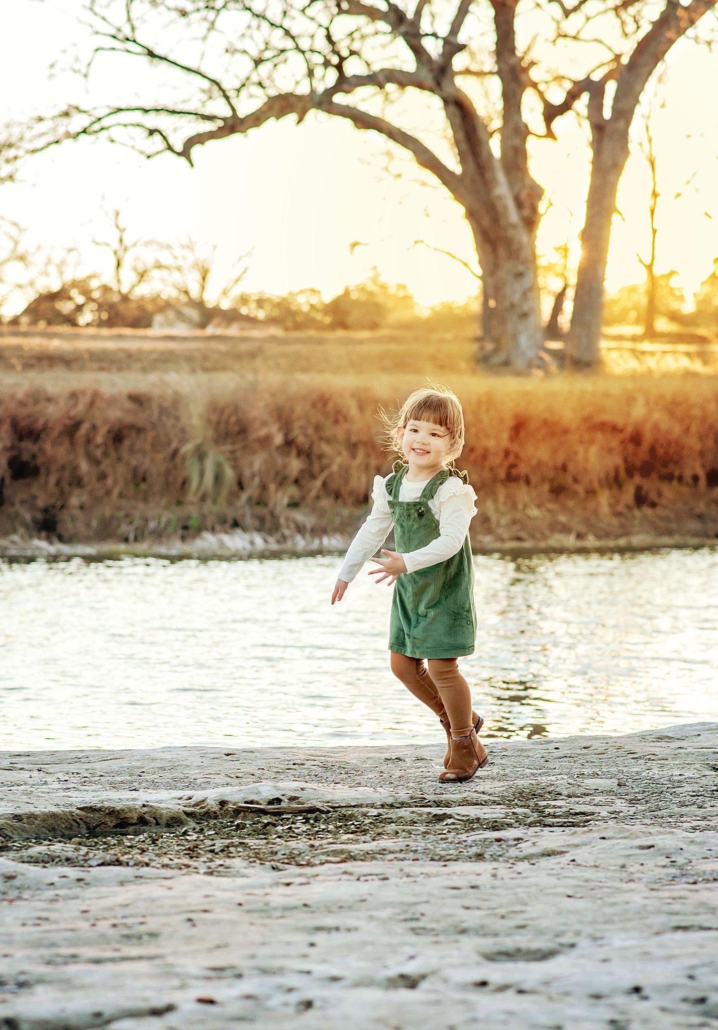 Small child in green vest runs and plays along a sandy beach at sunset with trees in background.