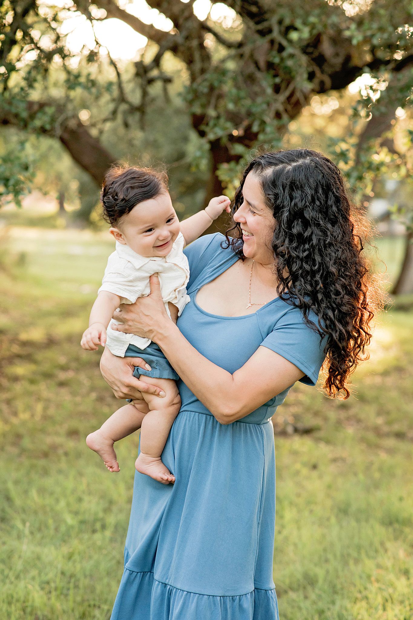 A parent in a blue dress holds a baby in white while laughing joyfully together outdoors in a sunny park setting.