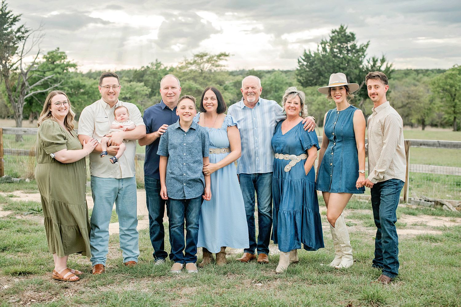 Large family group portrait outdoors on green grass, wearing coordinated blue and neutral tones, smiling together on a cloudy day.