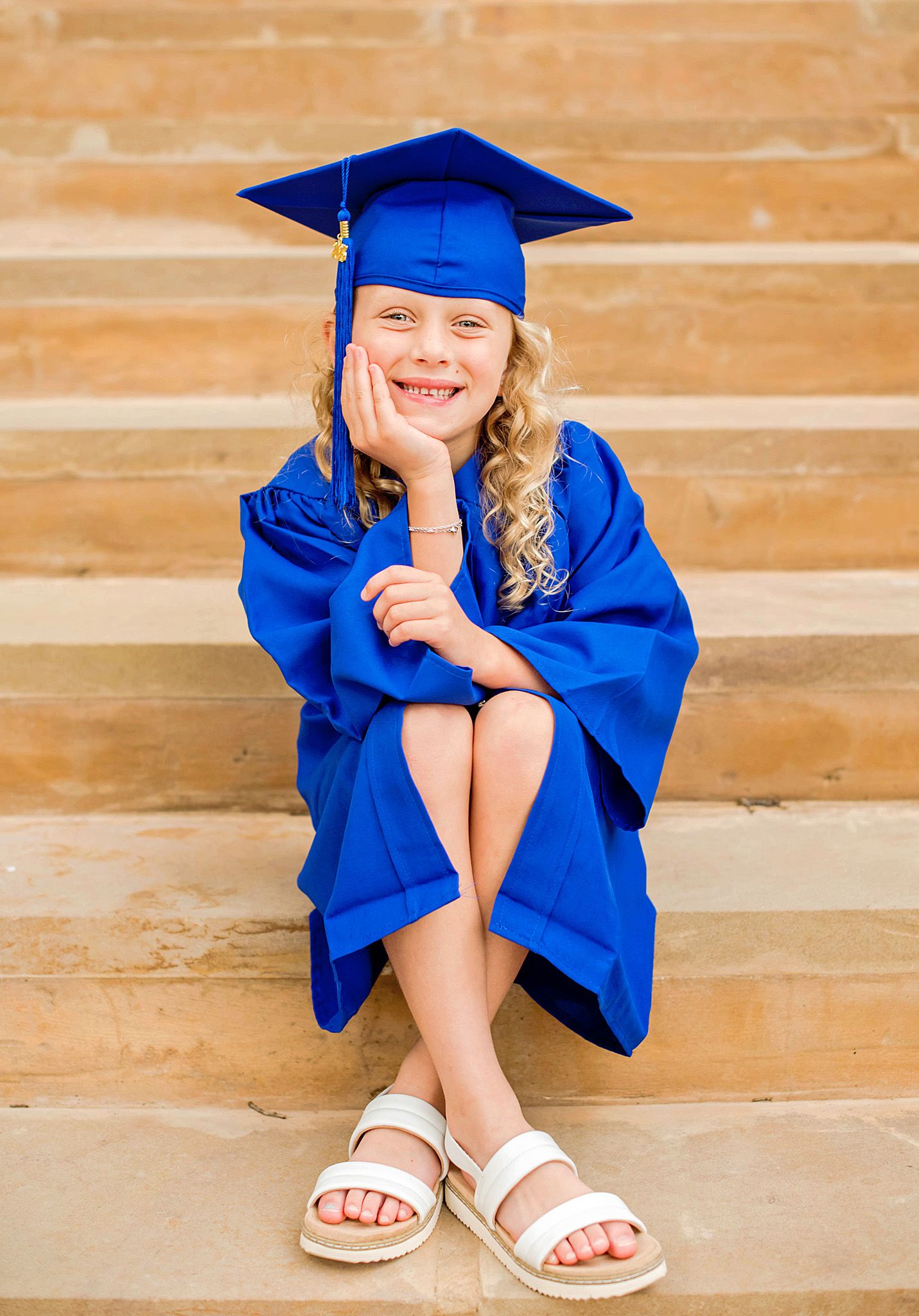 Young graduate in royal blue cap and gown sits on stone steps wearing white sandals during graduation photo session.