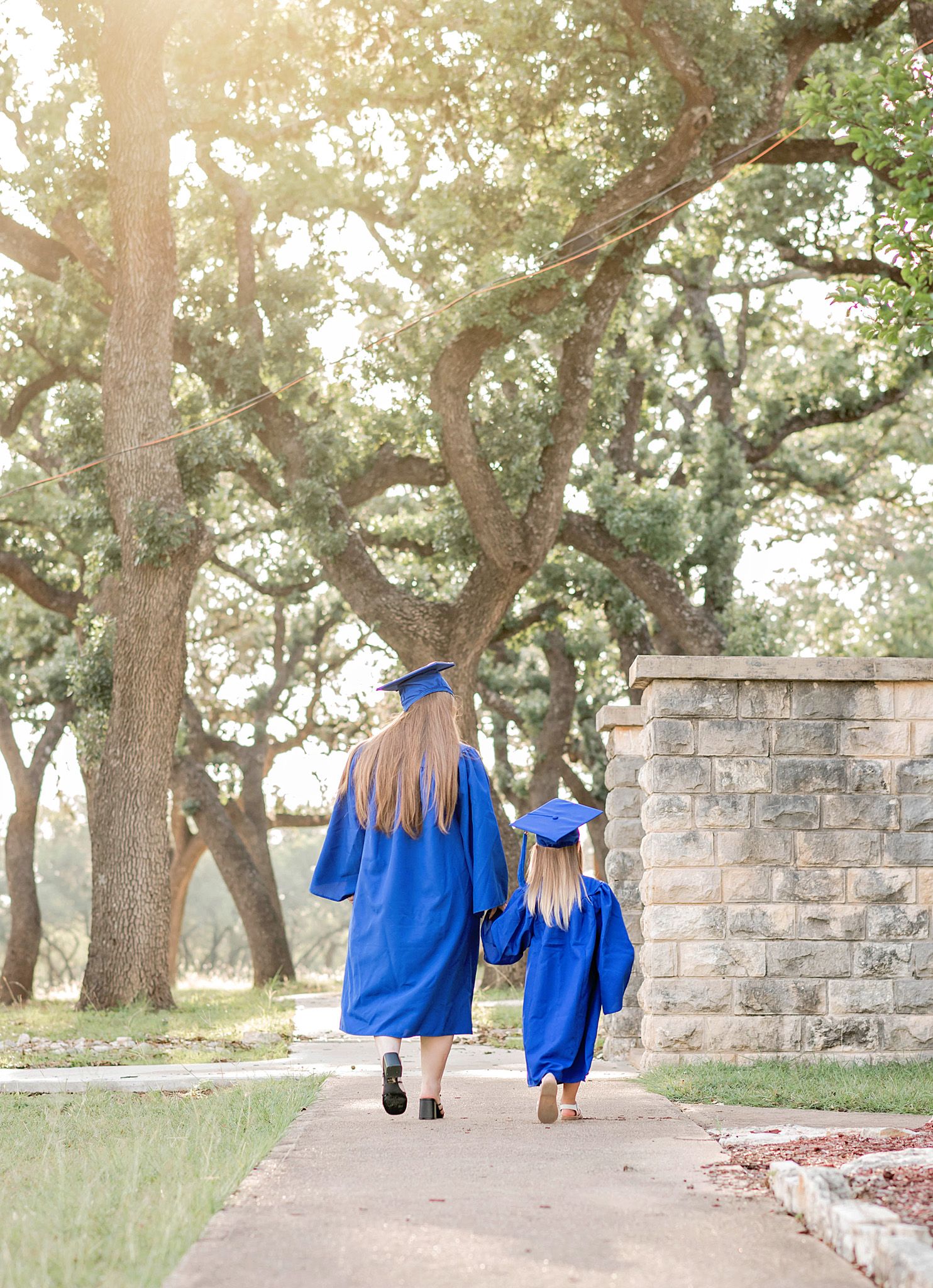 Two graduates in blue caps and gowns walk hand in hand down a sunlit path surrounded by oak trees.