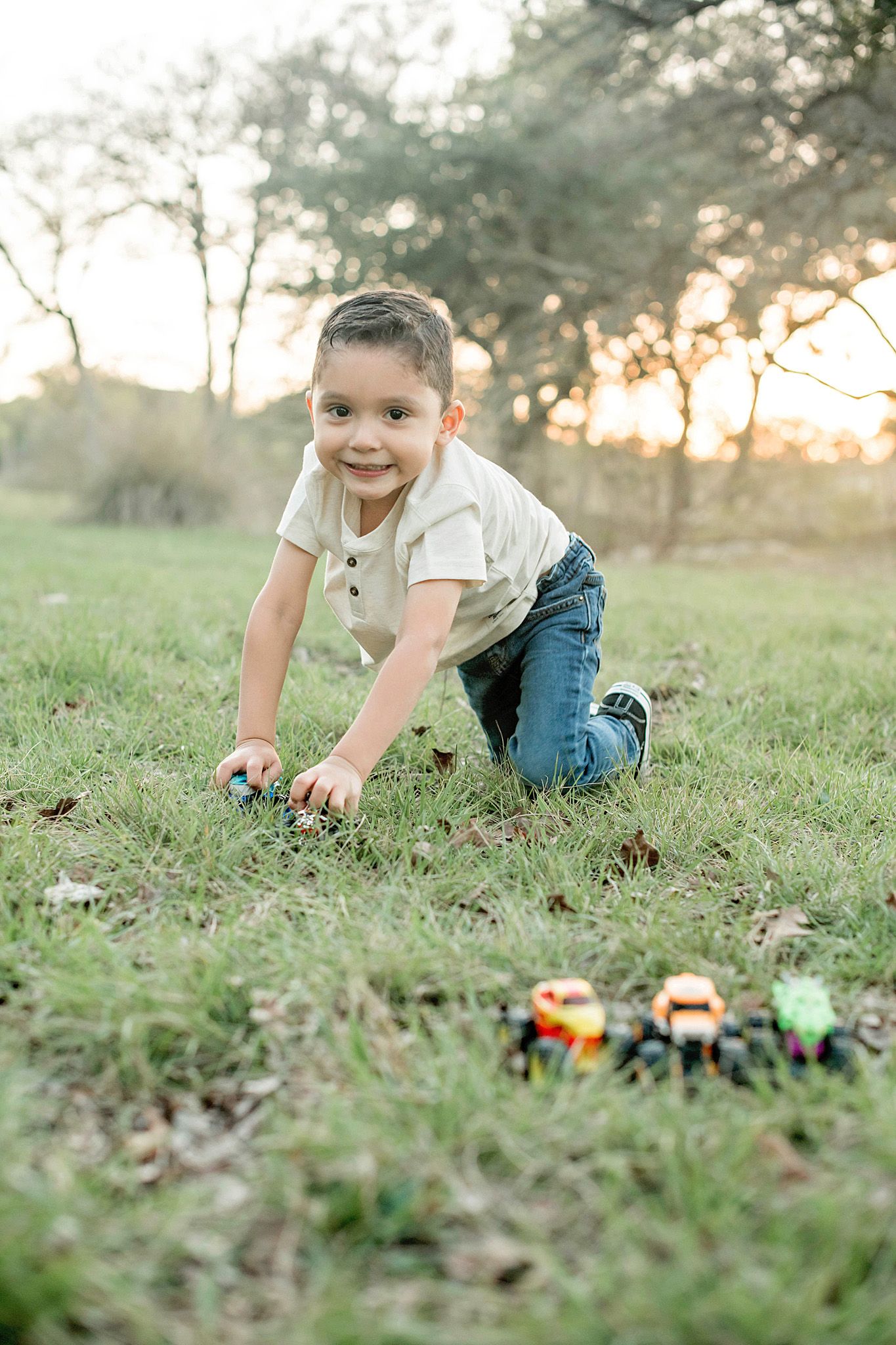 Young boy playing with toy cars on green grass at sunset, smiling at camera while kneeling outdoors.