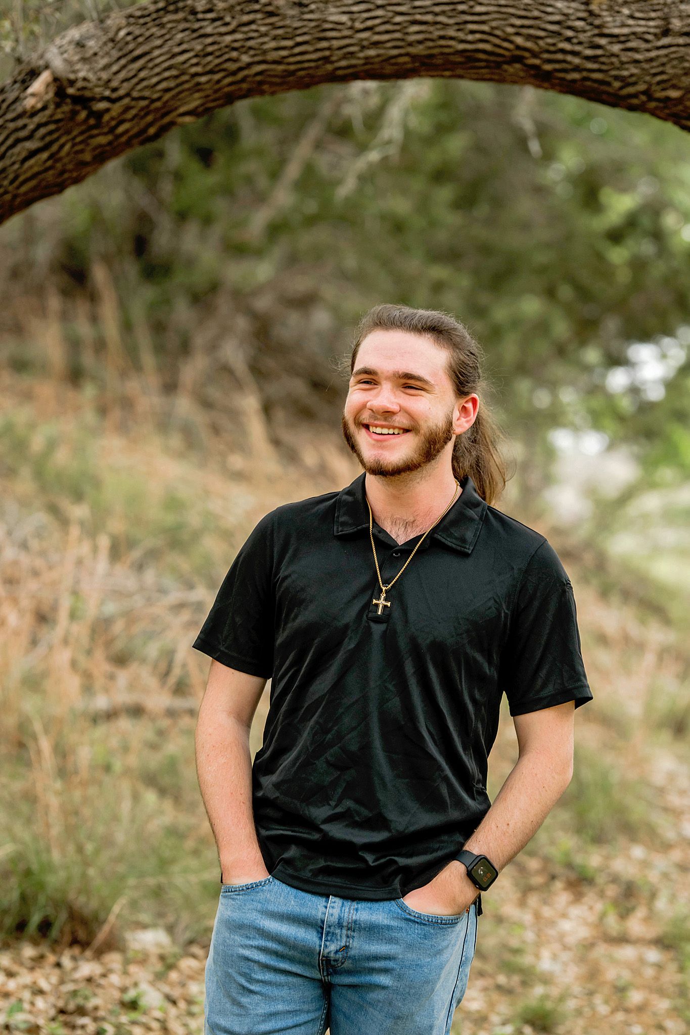 Person in black shirt and blue jeans poses casually outdoors against natural backdrop.