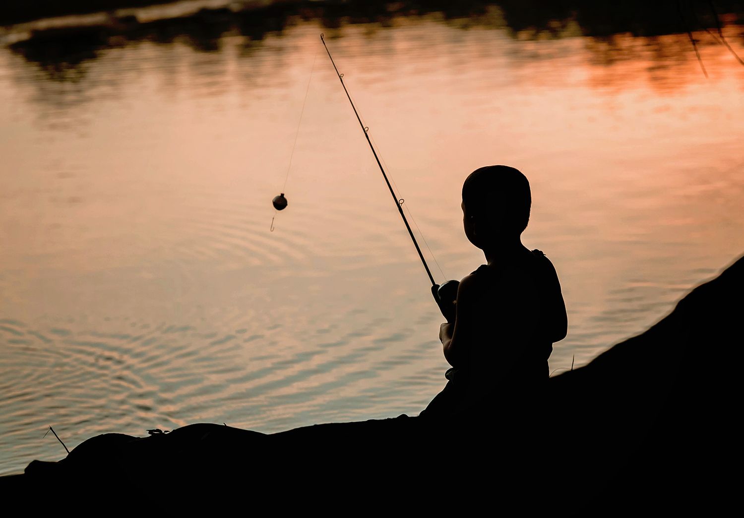 Silhouette of a young boy fishing at sunset by a calm lake with a fishing rod and line cast into the water.