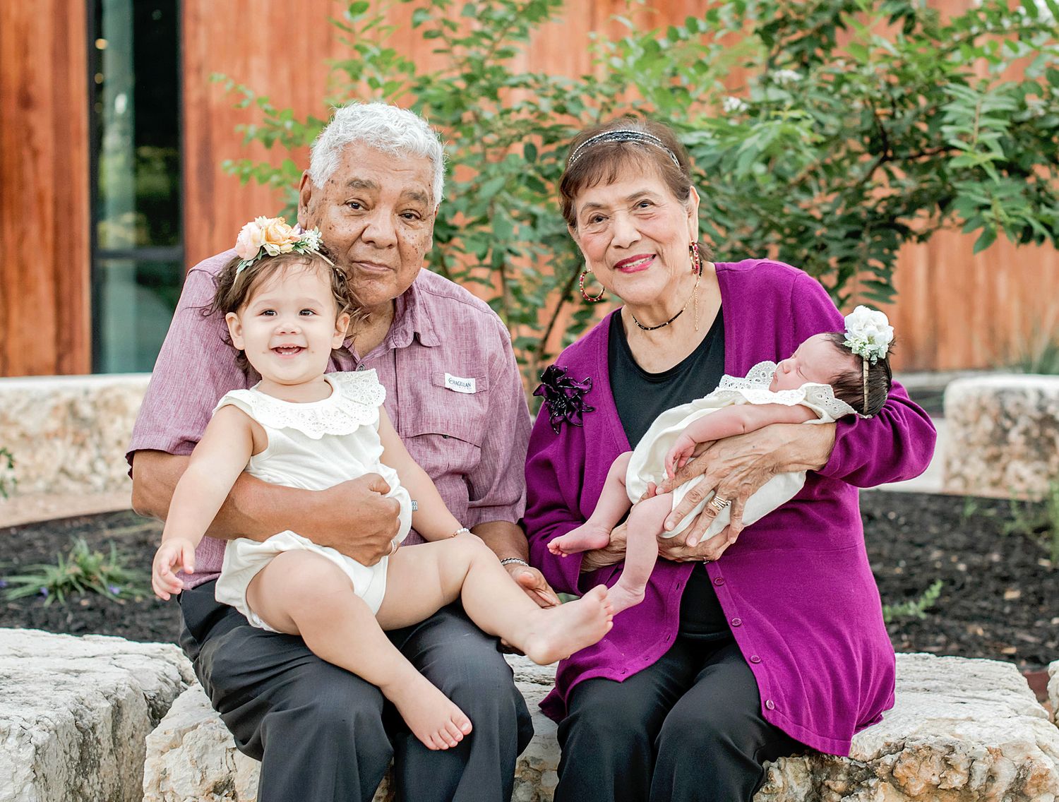 Grandparents sitting outdoors on stone bench, grandfather holding toddler girl, grandmother cradling newborn baby, smiling family portrait.